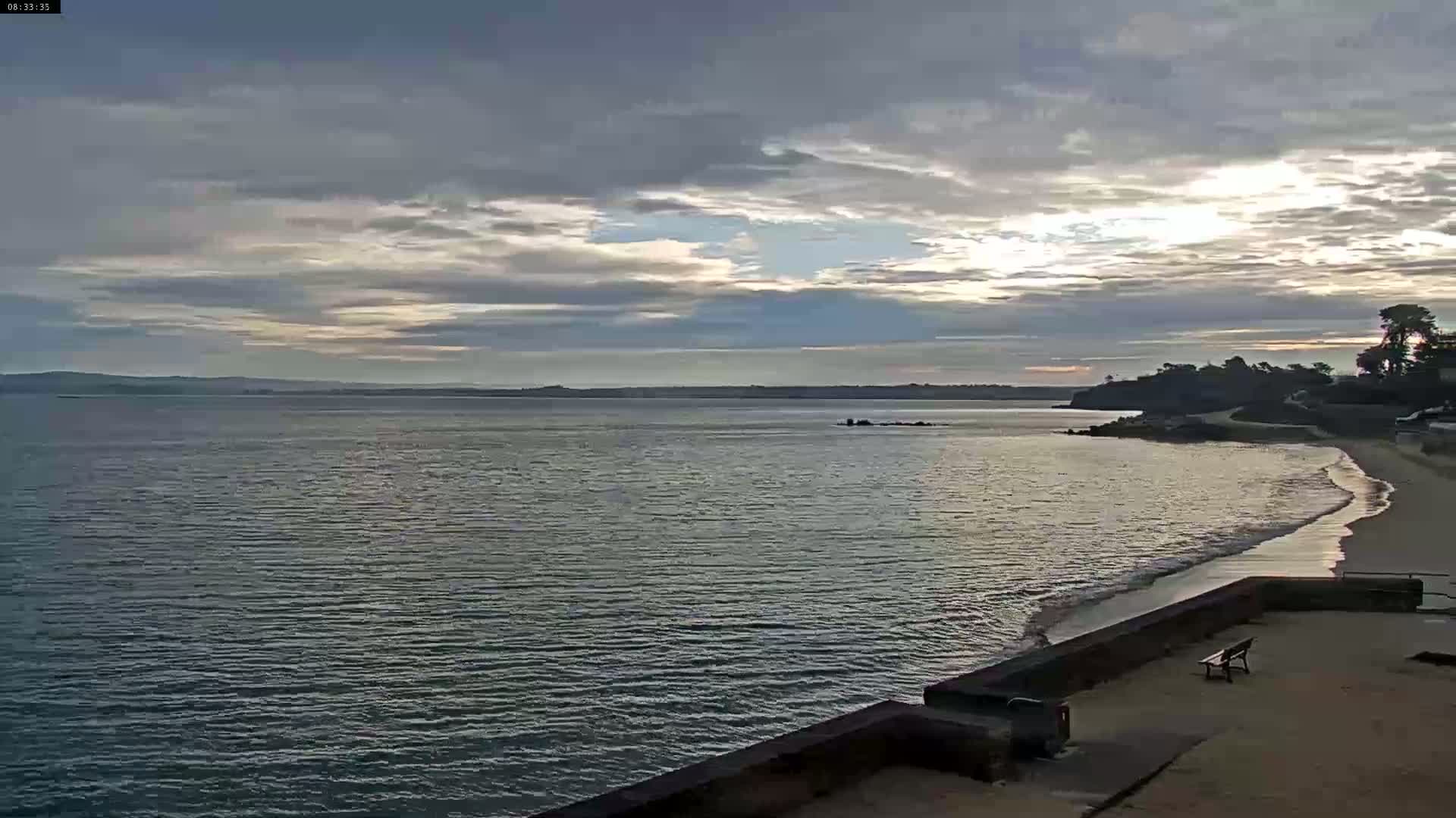 A calm body of water meets a sandy beach under a mostly cloudy sky with some patches of sunlight.