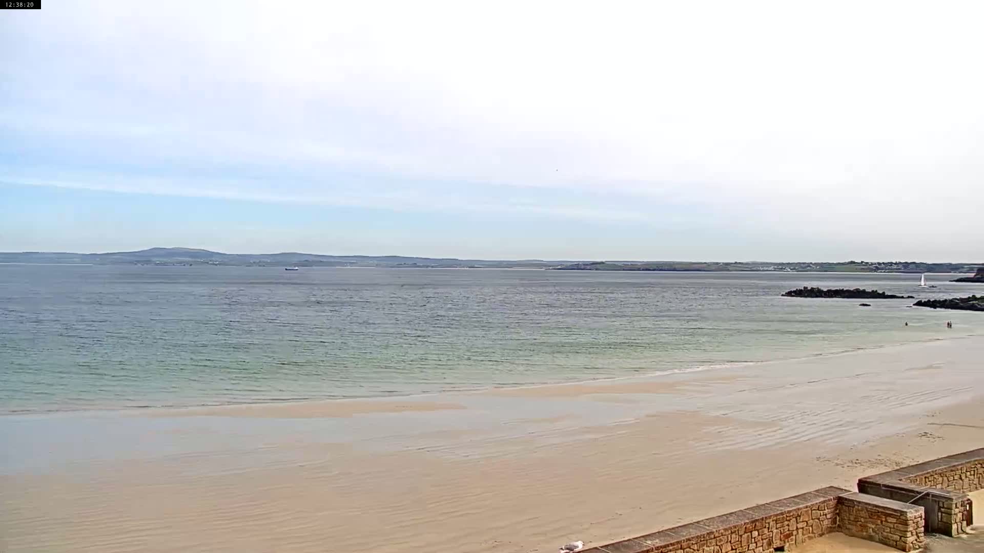 A wide sandy beach stretches to a calm sea under a mostly clear sky, with a distant landmass visible on the horizon.