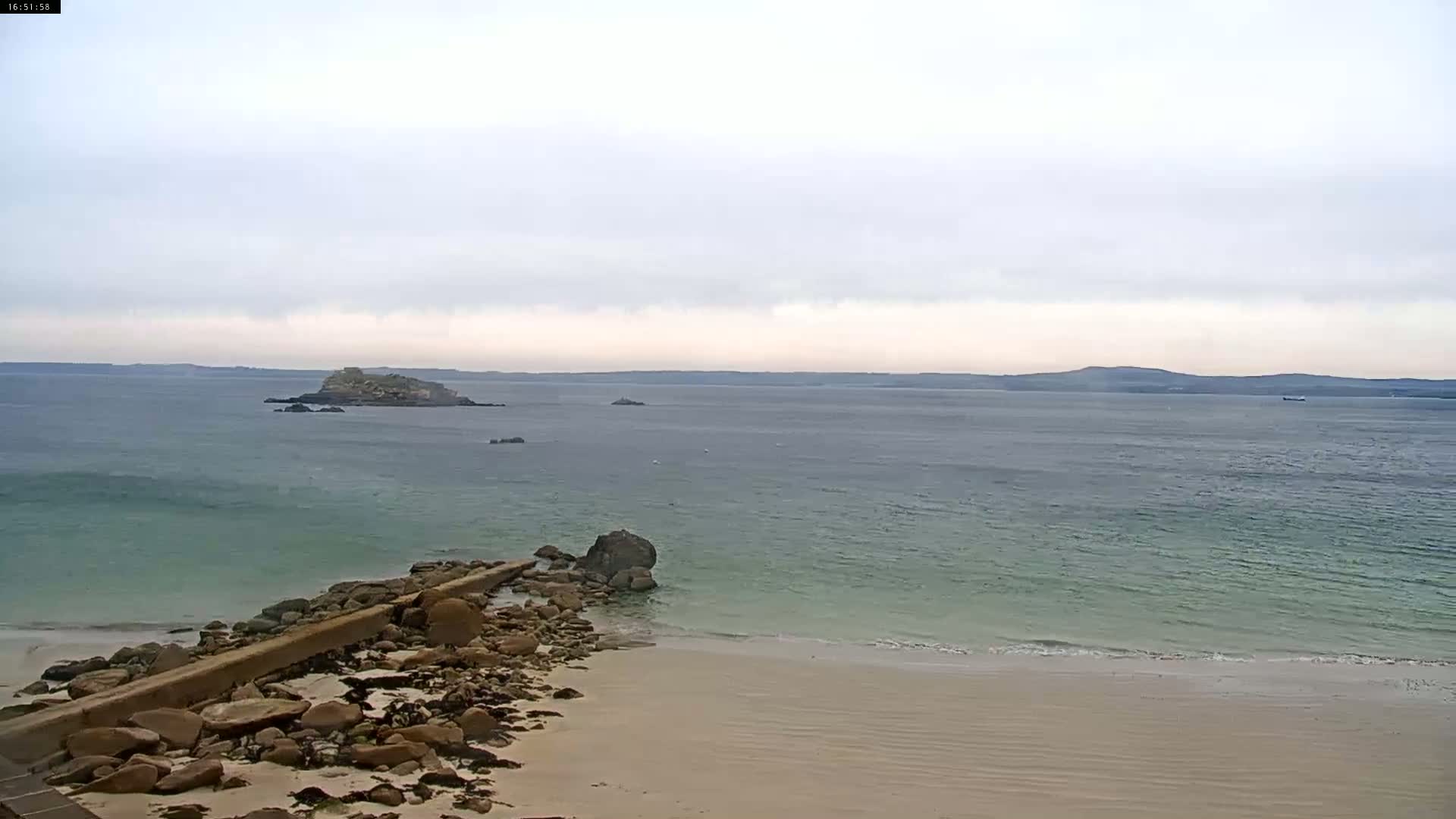 A rocky beach meets a calm, light-green ocean under an overcast sky, with an island visible in the distance.