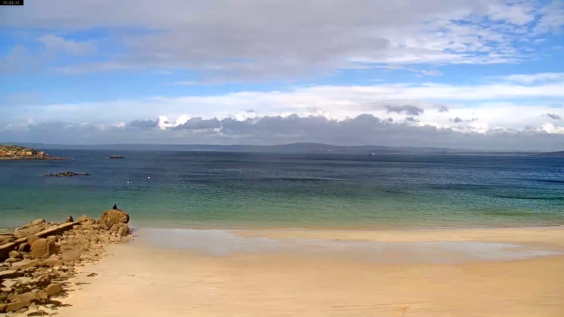 A sandy beach with calm, shallow, teal-colored water stretches to a dark blue ocean under a partly cloudy sky.