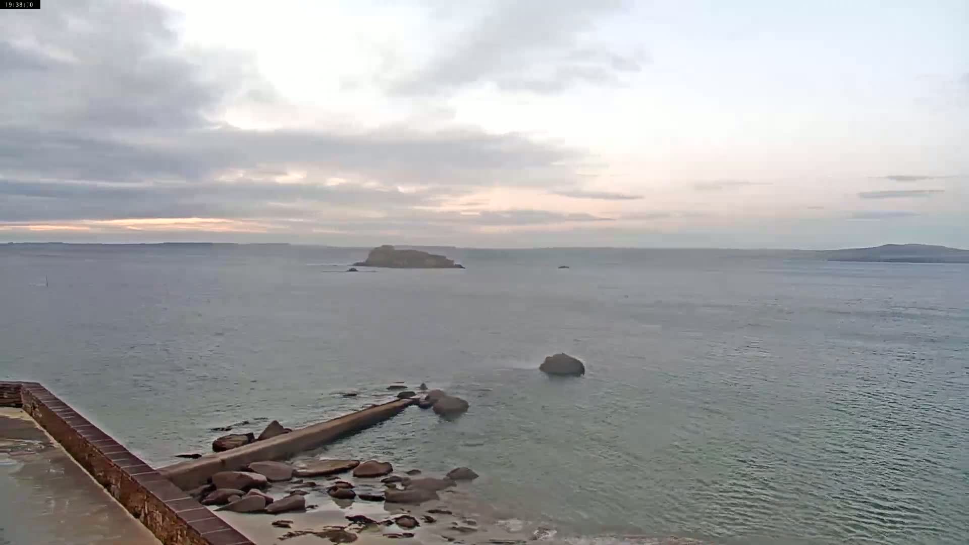A calm sea under a mostly cloudy sky at dusk shows a rocky shoreline with a low stone jetty extending into the water, and several small islands in the distance.