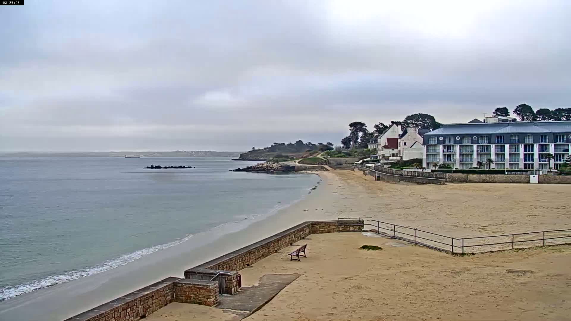 A sandy beach with gentle waves laps the shore under a cloudy sky, next to a multi-story hotel and other buildings.