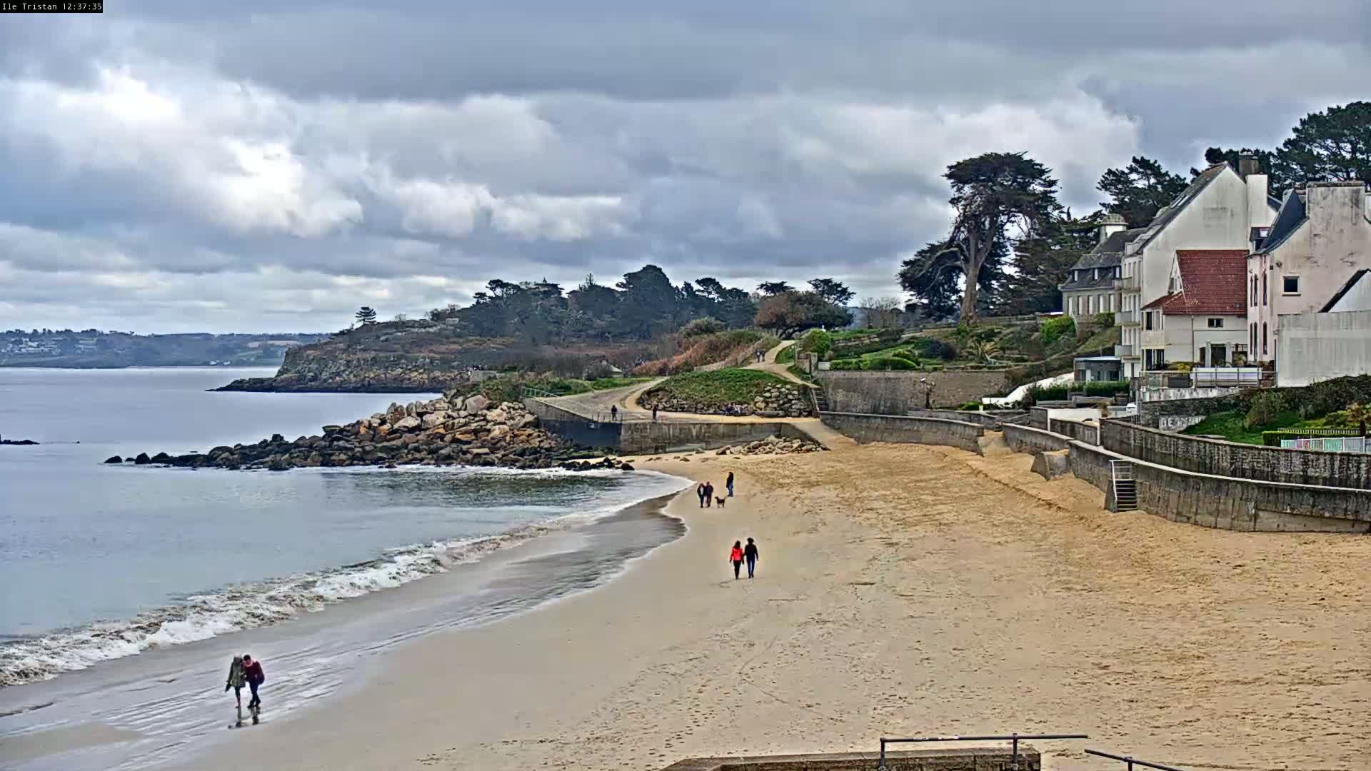 A sandy beach with several people walking along it, overlooks a calm bay under a cloudy sky, with white buildings perched on a cliff in the background.