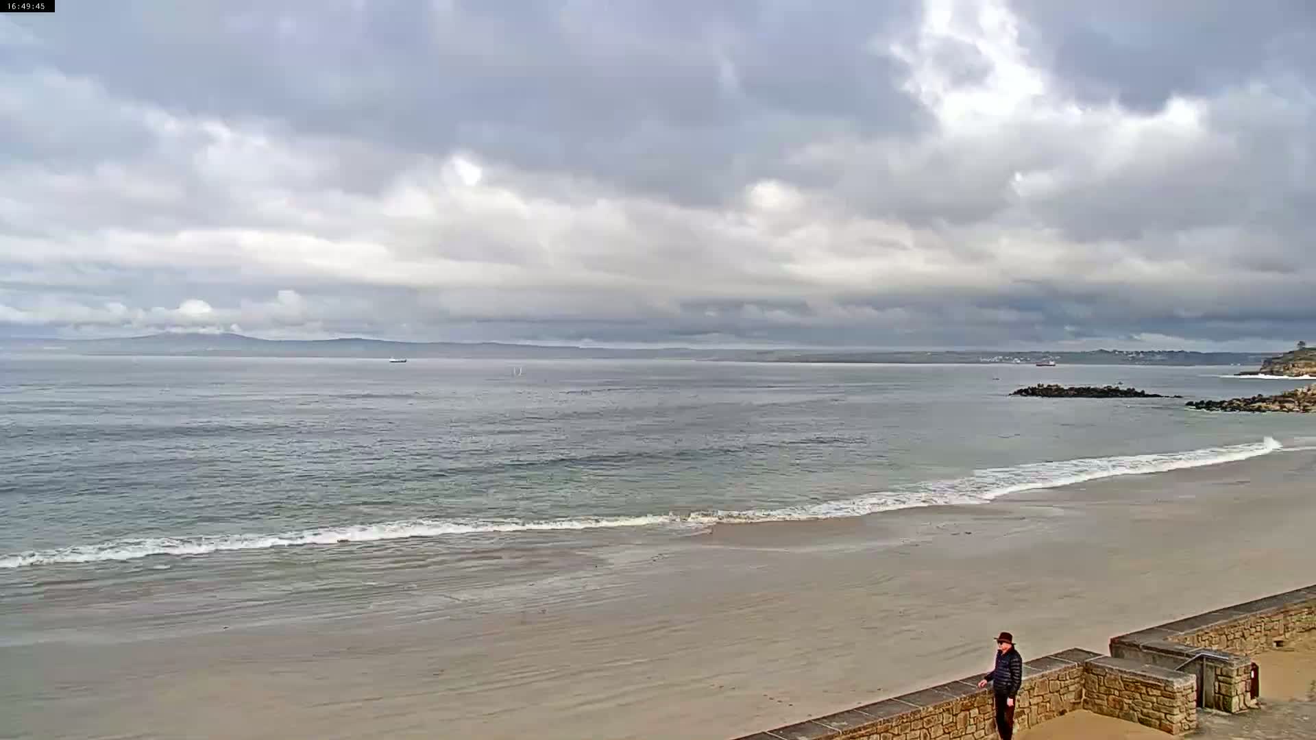 A person stands on a stone wall overlooking a calm, grey ocean with a sandy beach under a cloudy sky.