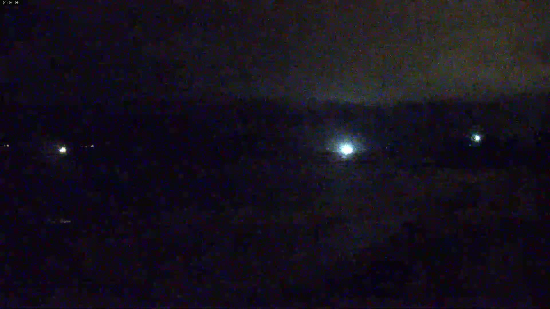 A nighttime, dark scene shows a few faint lights in the distance under a mostly cloudy sky.