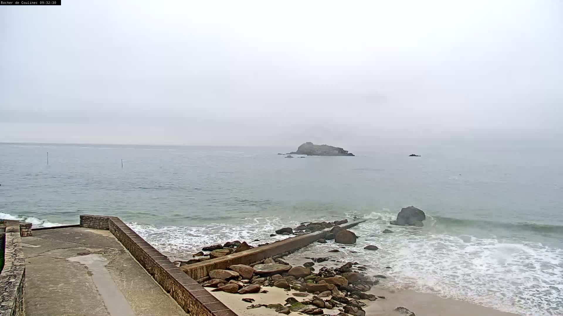 A rocky shoreline with a concrete structure meets a misty ocean where a small island is visible in the distance.
