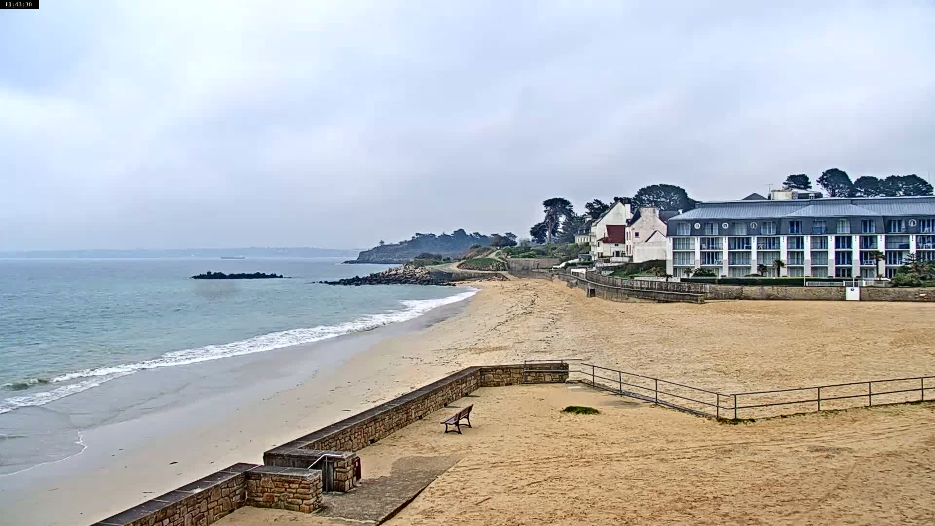 A sandy beach with a low stone wall and a multi-story building overlooks a calm ocean under an overcast sky.