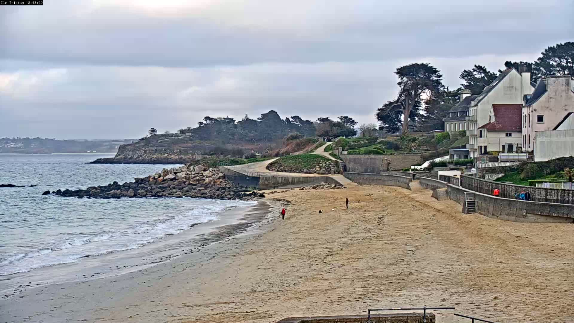 A sandy beach with gentle waves laps the shore under a cloudy sky,  with a few people walking along the beach and several houses perched on a cliff overlooking the water.