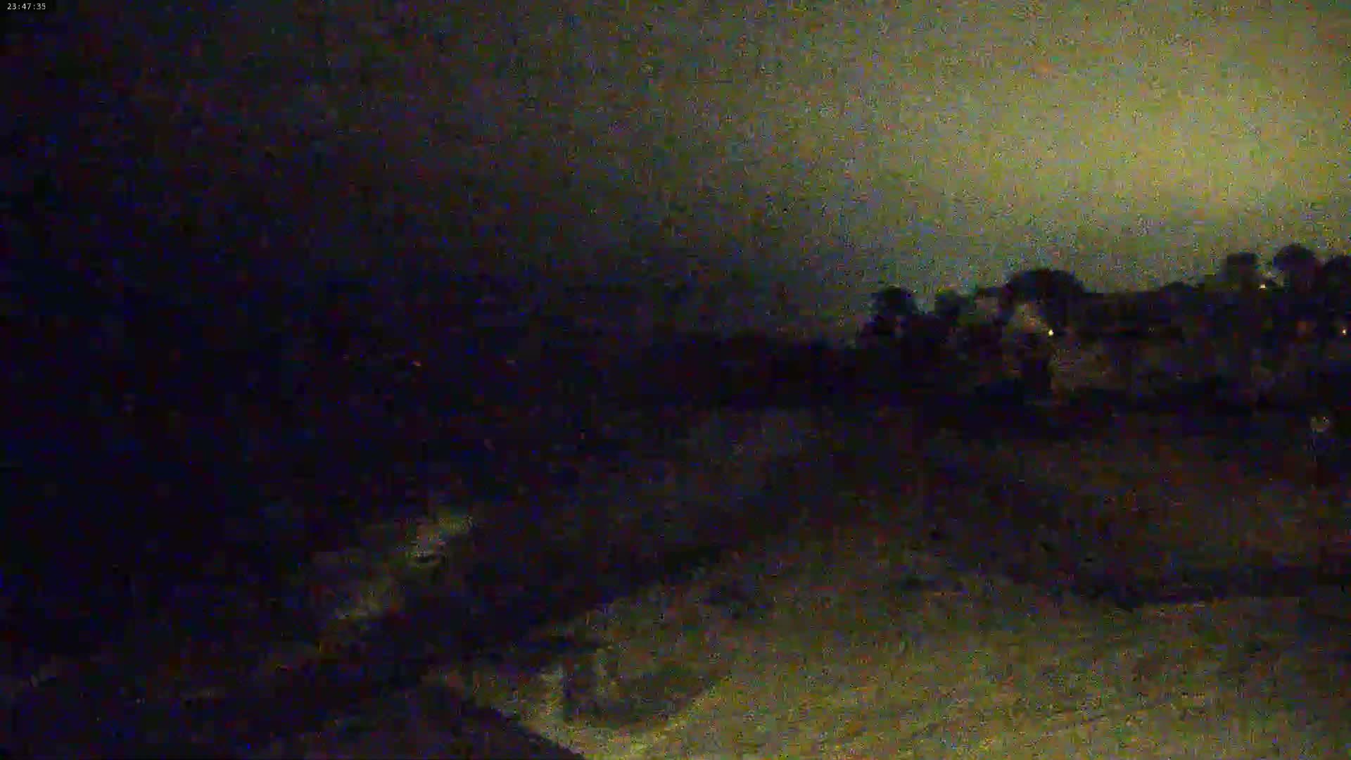A dark, nighttime scene shows a sparsely lit area with trees and some faint lights in the distance under a mostly cloudy sky.