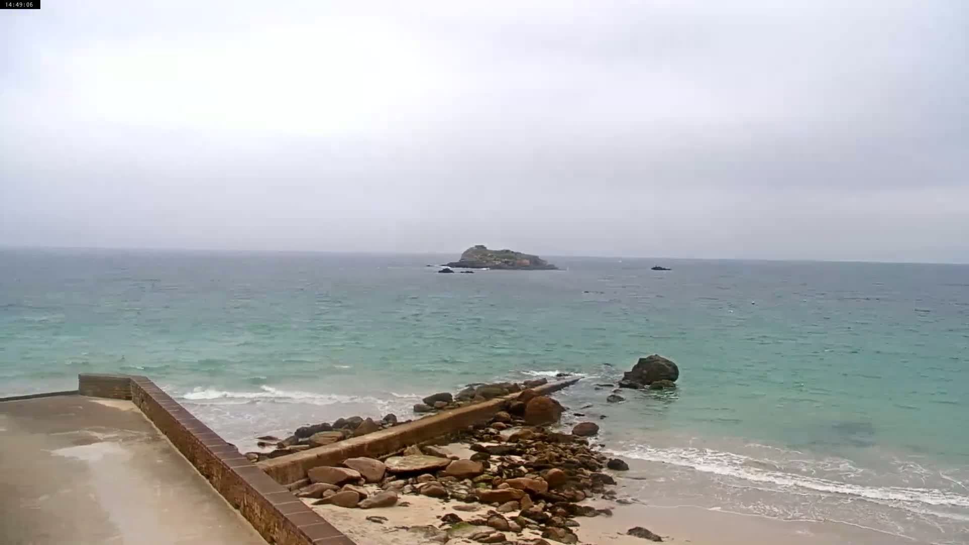 A rocky shoreline with a low concrete wall meets a teal ocean with small waves under an overcast sky, and a small island is visible in the distance.