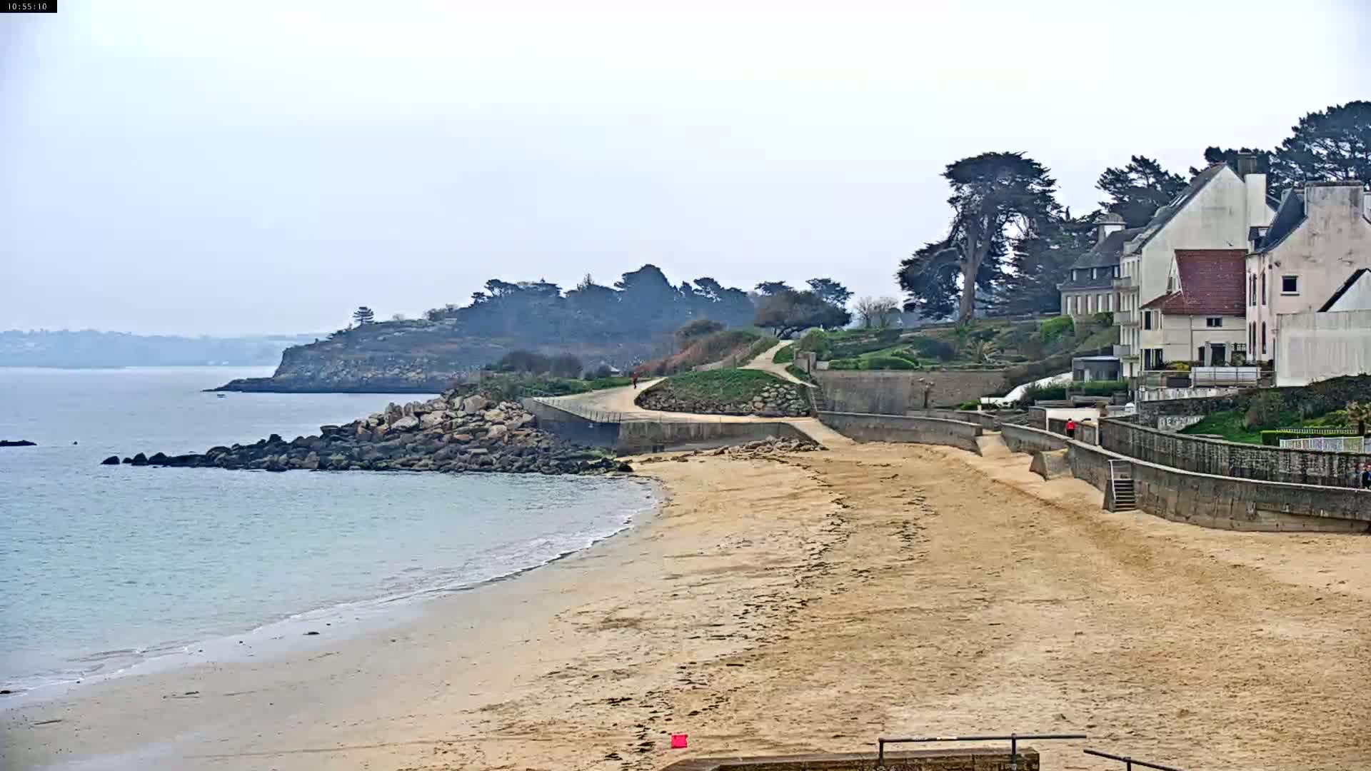 A sandy beach curves gently toward a calm body of water, bordered by a rocky shoreline and a line of houses perched on a hillside under an overcast sky.