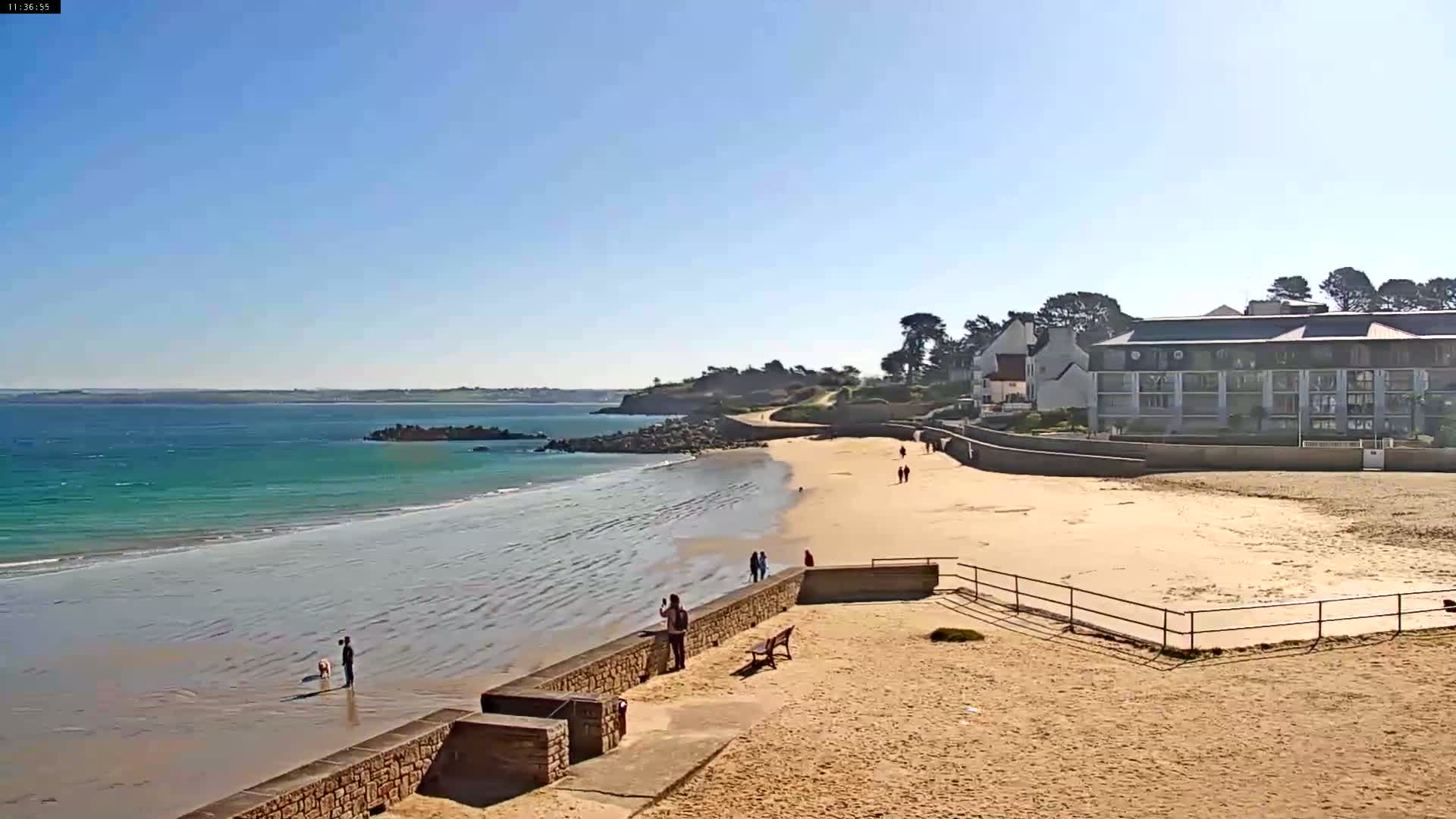 A sandy beach with calm, turquoise water stretches out under a clear blue sky, with a few people strolling along the shore and a multi-story building visible in the background.