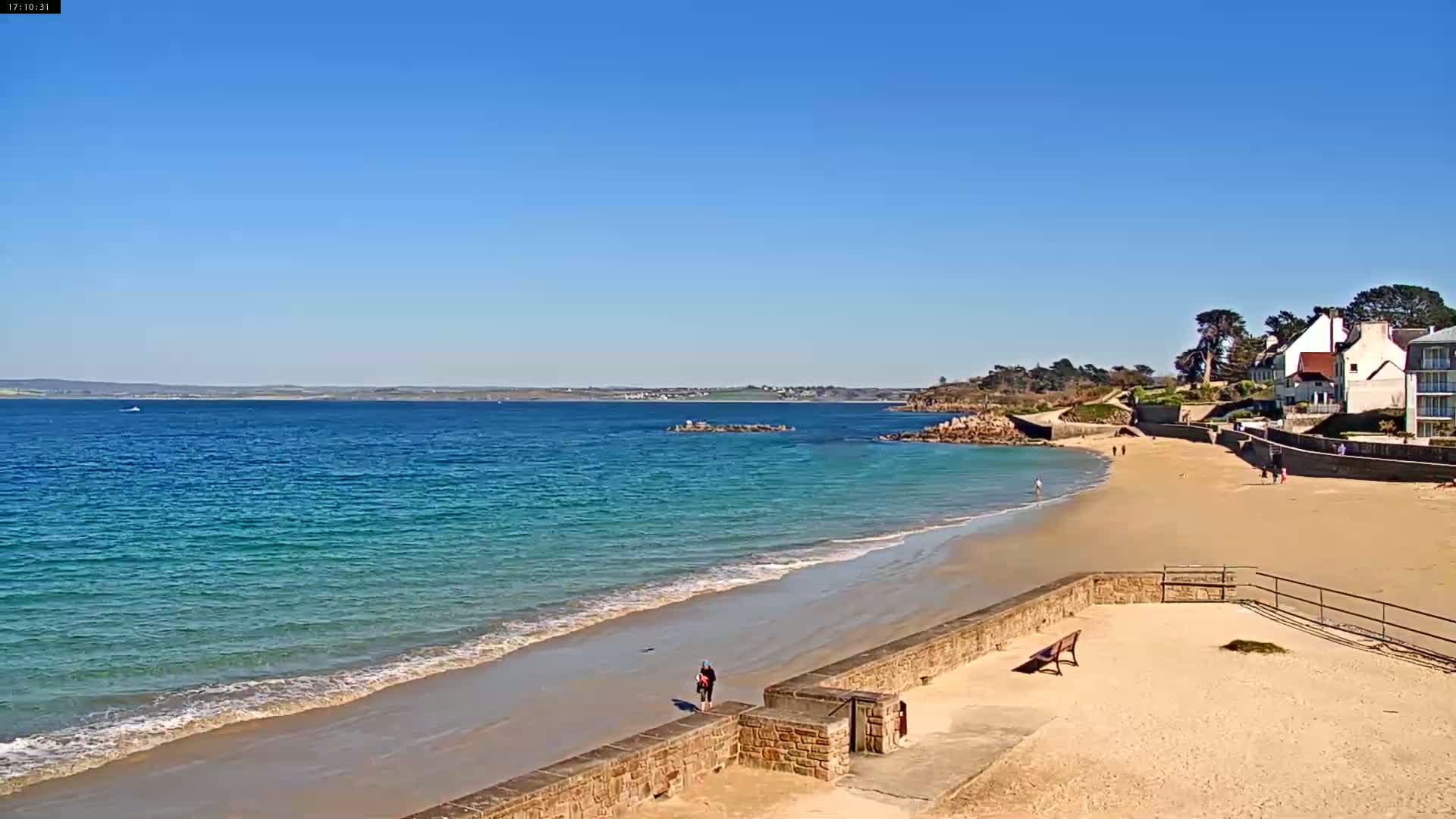 A sandy beach with calm, turquoise water stretches out under a clear blue sky, next to a low stone wall and several buildings.