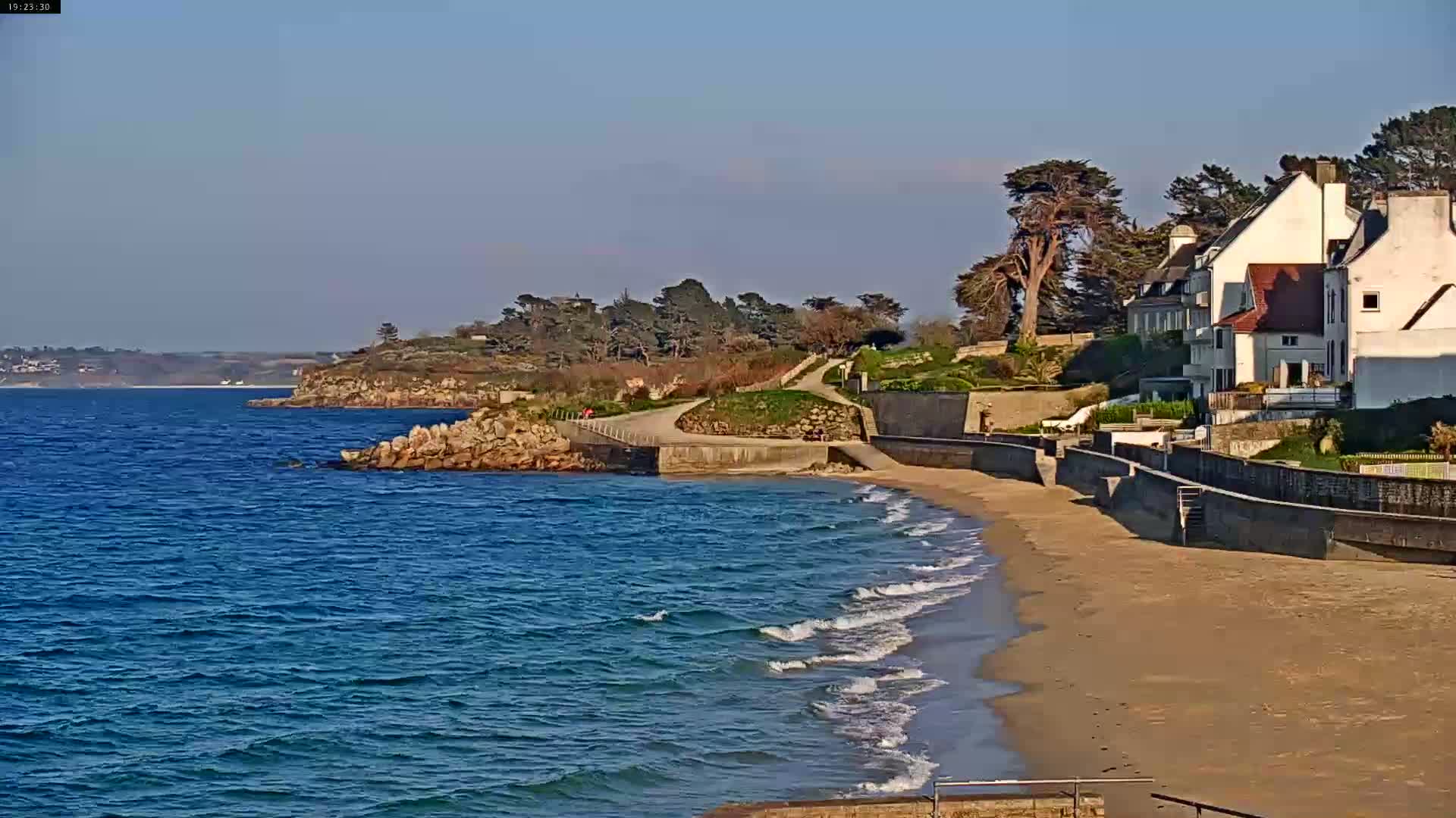 A sandy beach with small waves laps the shore near a paved walkway leading to several houses on a coastal cliff under a clear, sunny sky.