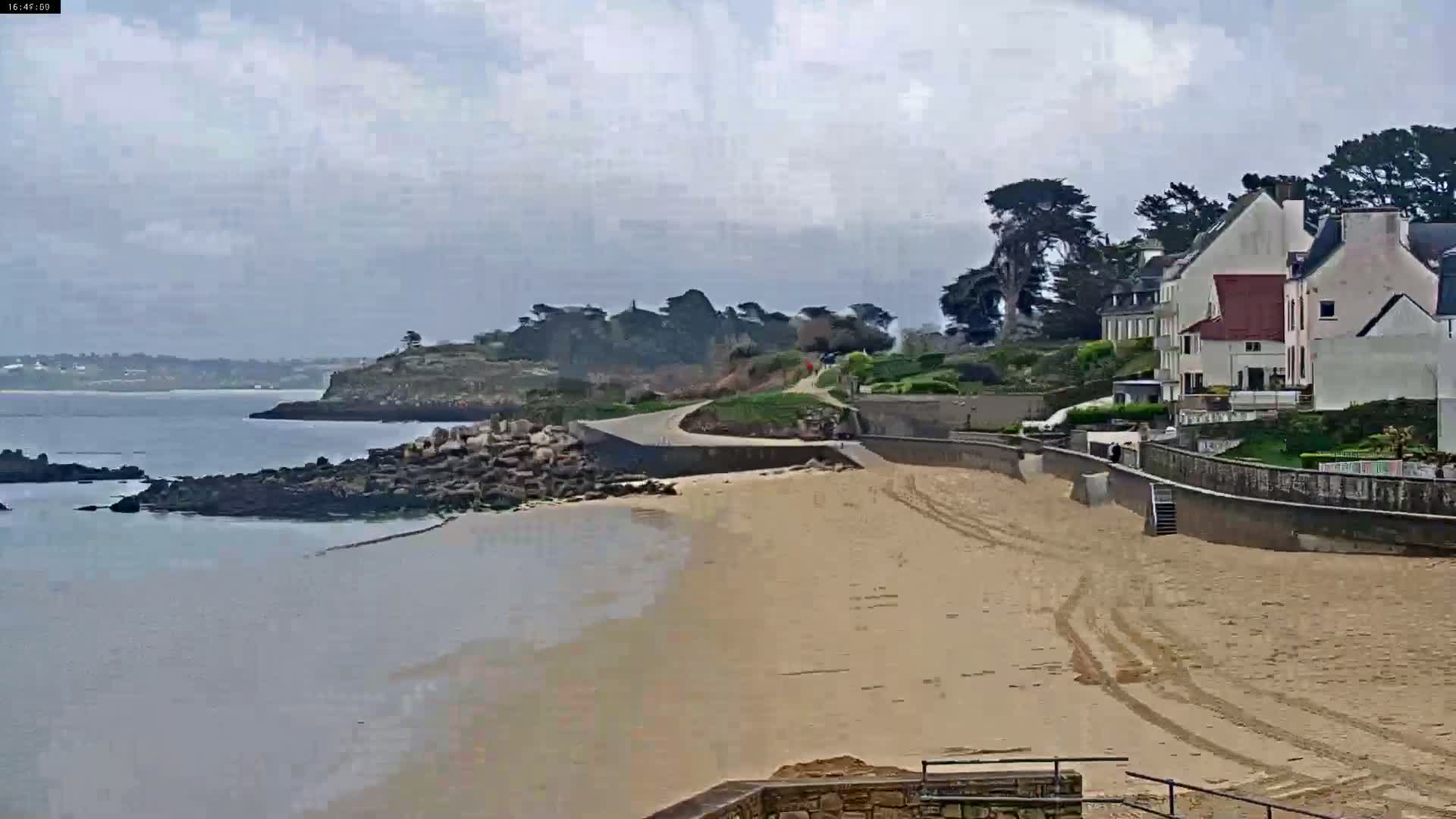 A sandy beach curves around a rocky shoreline under a cloudy sky, with houses and greenery situated on a hillside overlooking the water.