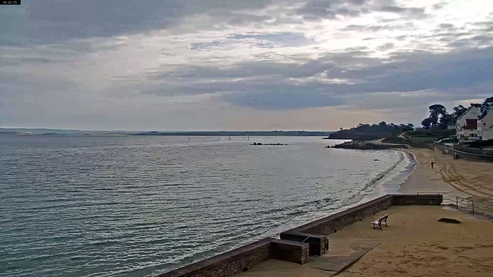 A calm, cloudy day at a beach with small waves lapping a sandy shore, a stone wall, and a few buildings in the distance.