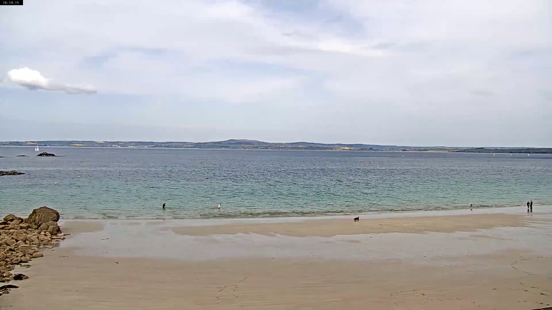 A sandy beach stretches to a calm, pale-blue sea under a mostly cloudy sky, with a few people and a dog visible on the shore and distant land visible across the water.