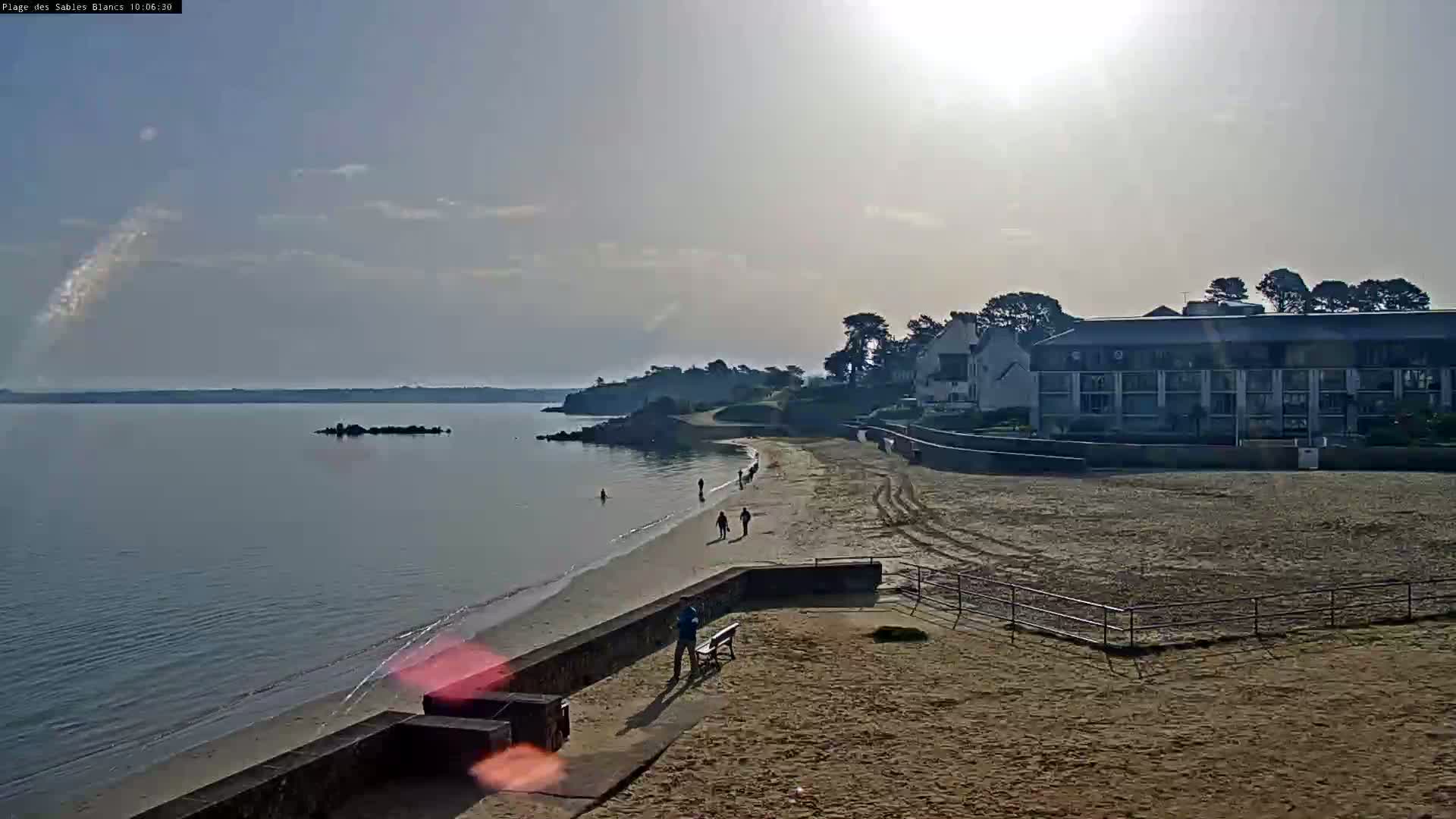 A sandy beach with a few people walking along the shoreline, a calm body of water, and a large building in the background under a mostly clear, sunny sky.