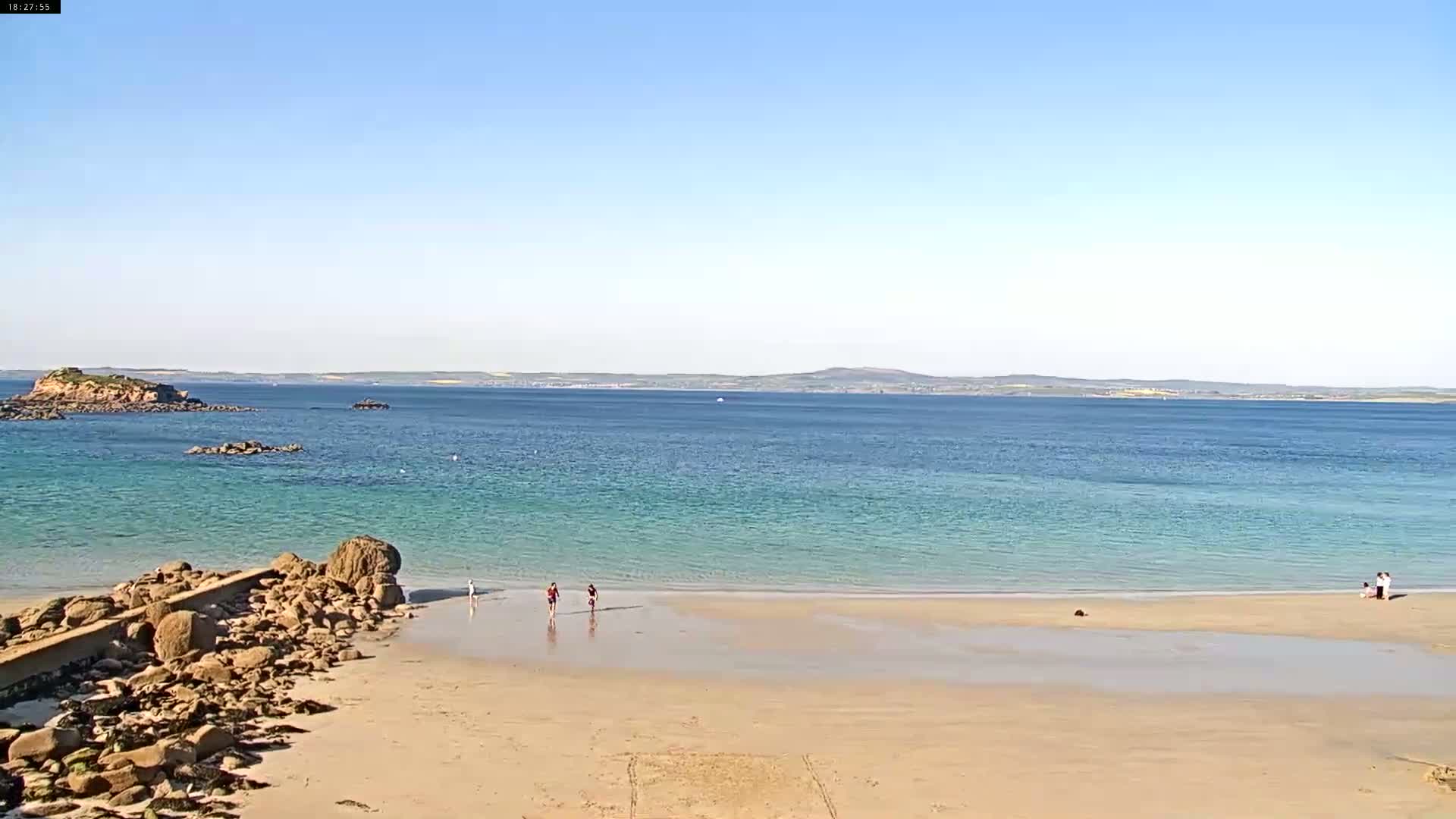 A sandy beach with a few people on it slopes gently to a calm, light teal ocean under a clear blue sky, with a small island visible in the distance.