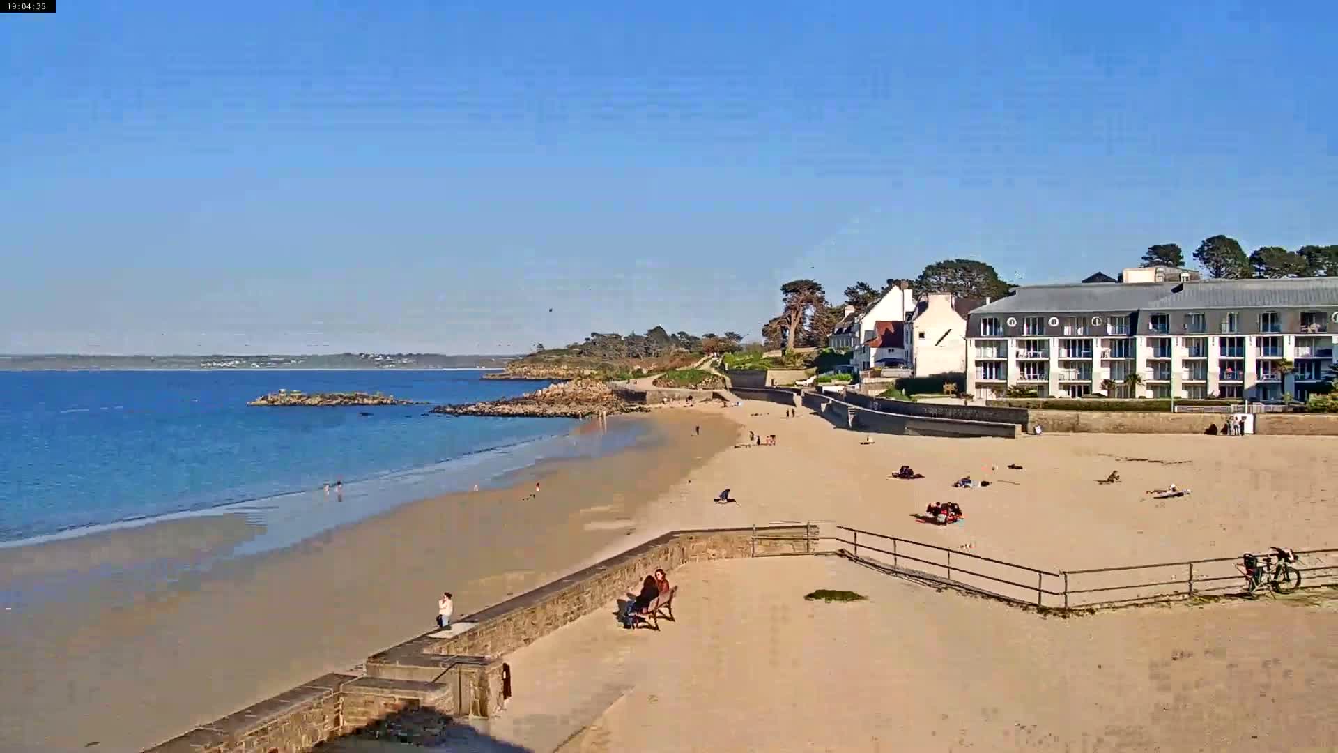 A sandy beach with a calm ocean and several people enjoying a sunny day, with buildings situated along the shoreline.