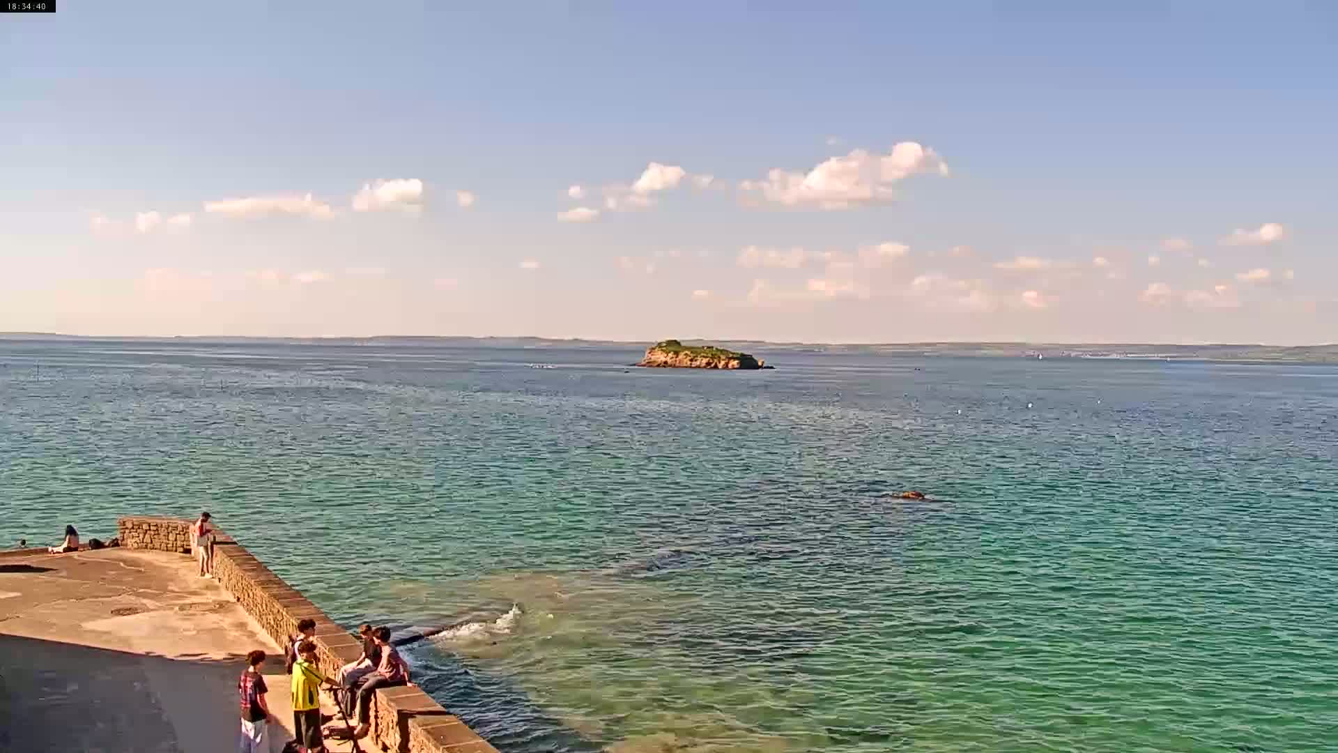 A sunny day shows a calm, teal-colored body of water with a small island in the distance and several people gathered on a stone wall near the water's edge.