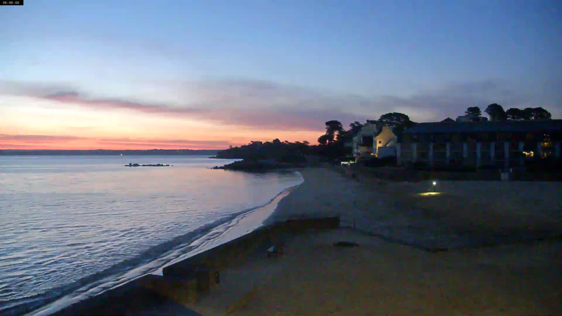 A calm body of water meets a sandy beach at sunset, with a building and trees silhouetted against the colorful sky.