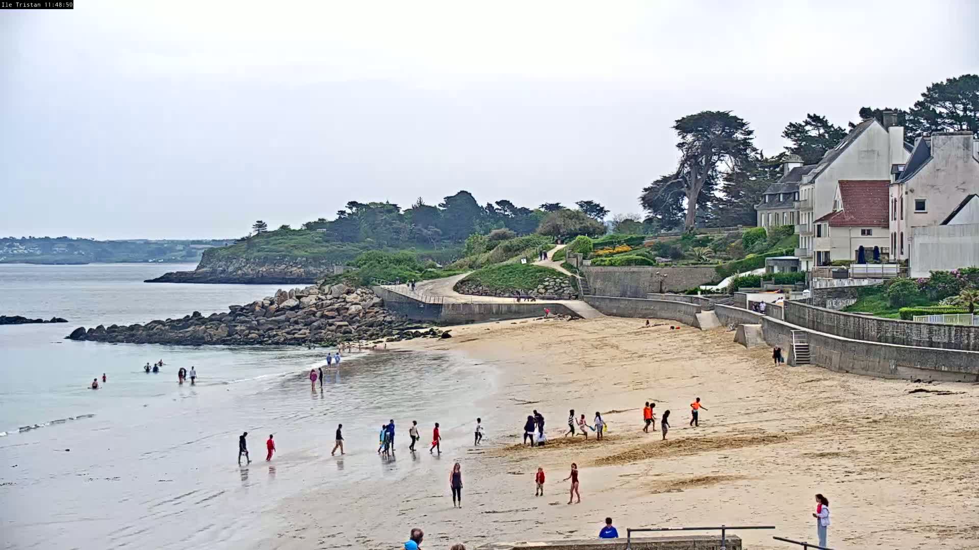 A sandy beach with several people on it, some in the water, and a calm bay backed by a rocky shoreline and houses, under an overcast sky.