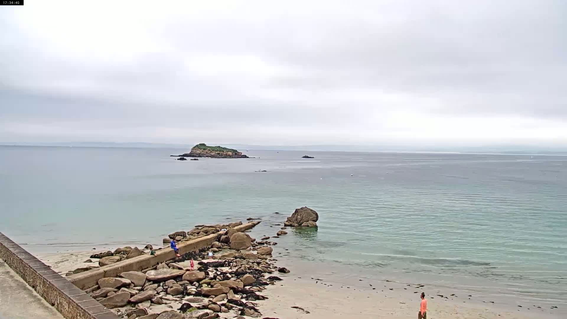 A calm, overcast day at a beach with a low stone wall extending into the water, a small island visible in the distance, and several people on the rocks and sand.