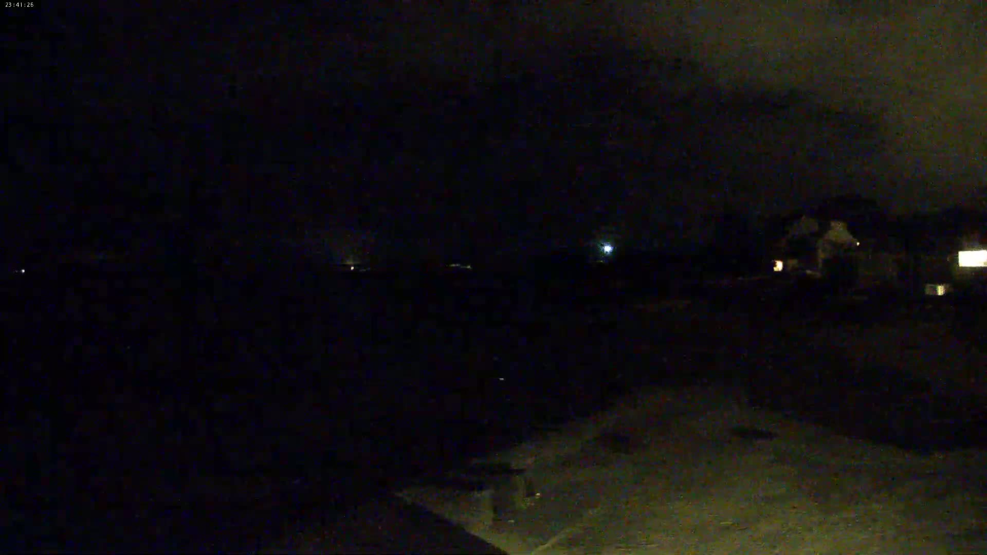 A nighttime, dark outdoor scene shows a few dimly lit houses and a mostly obscured landscape under a cloudy sky.