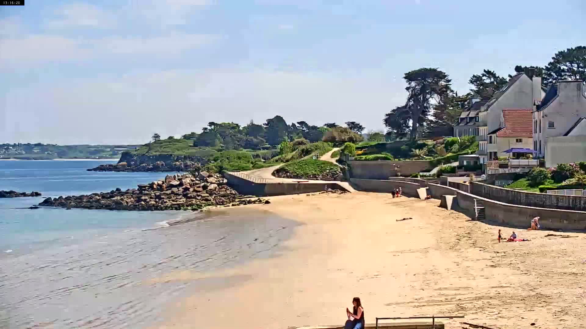 A sandy beach stretches out to a calm sea, bordered by rocky shores and a line of houses overlooking the water under a sunny sky.