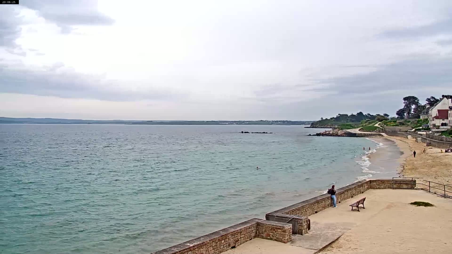 A sandy beach with calm, light-blue water stretches out to a distant coastline under a cloudy sky.