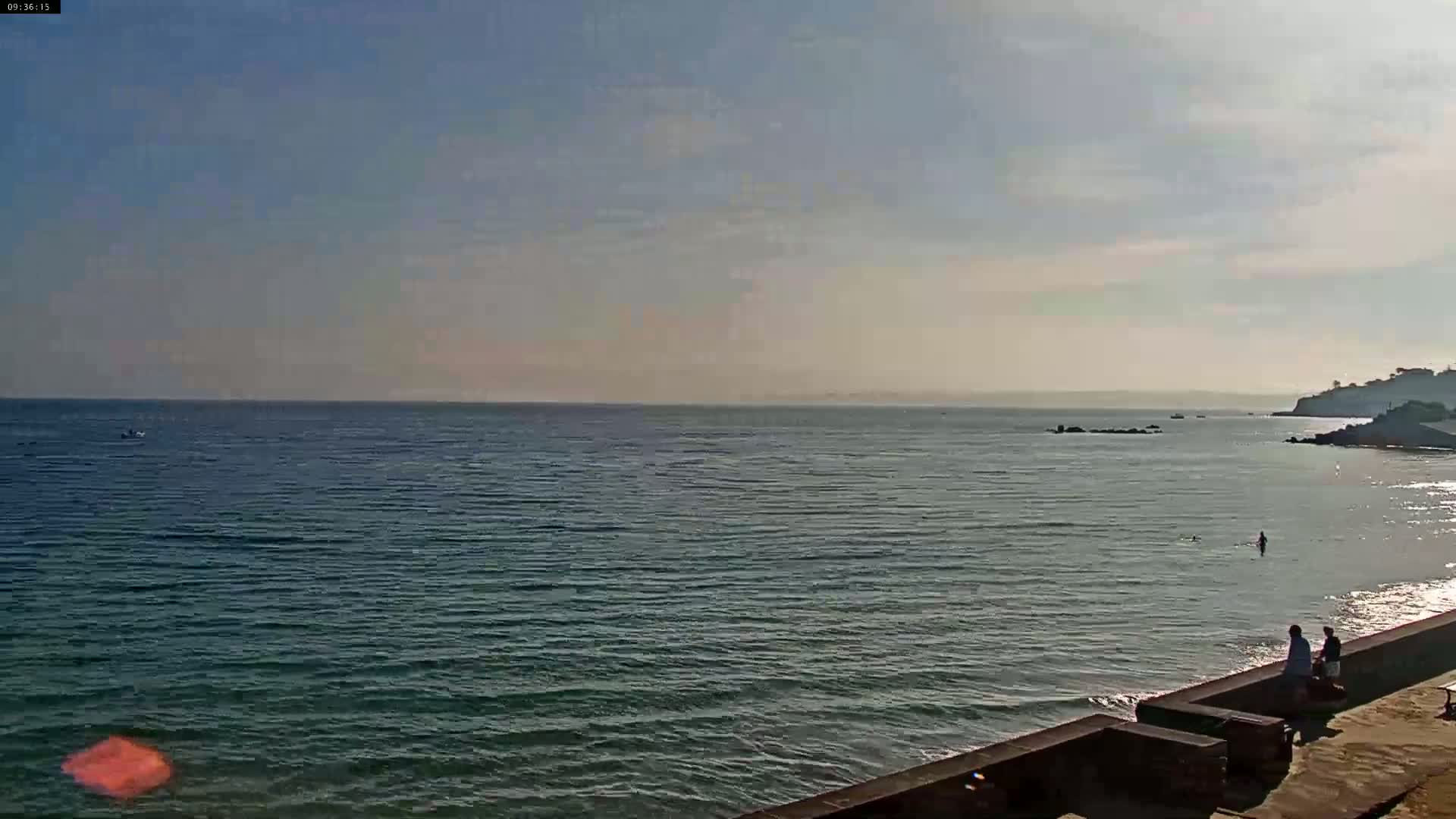 A calm, sunny ocean scene shows a few people near a low wall overlooking a tranquil body of water with a distant coastline.