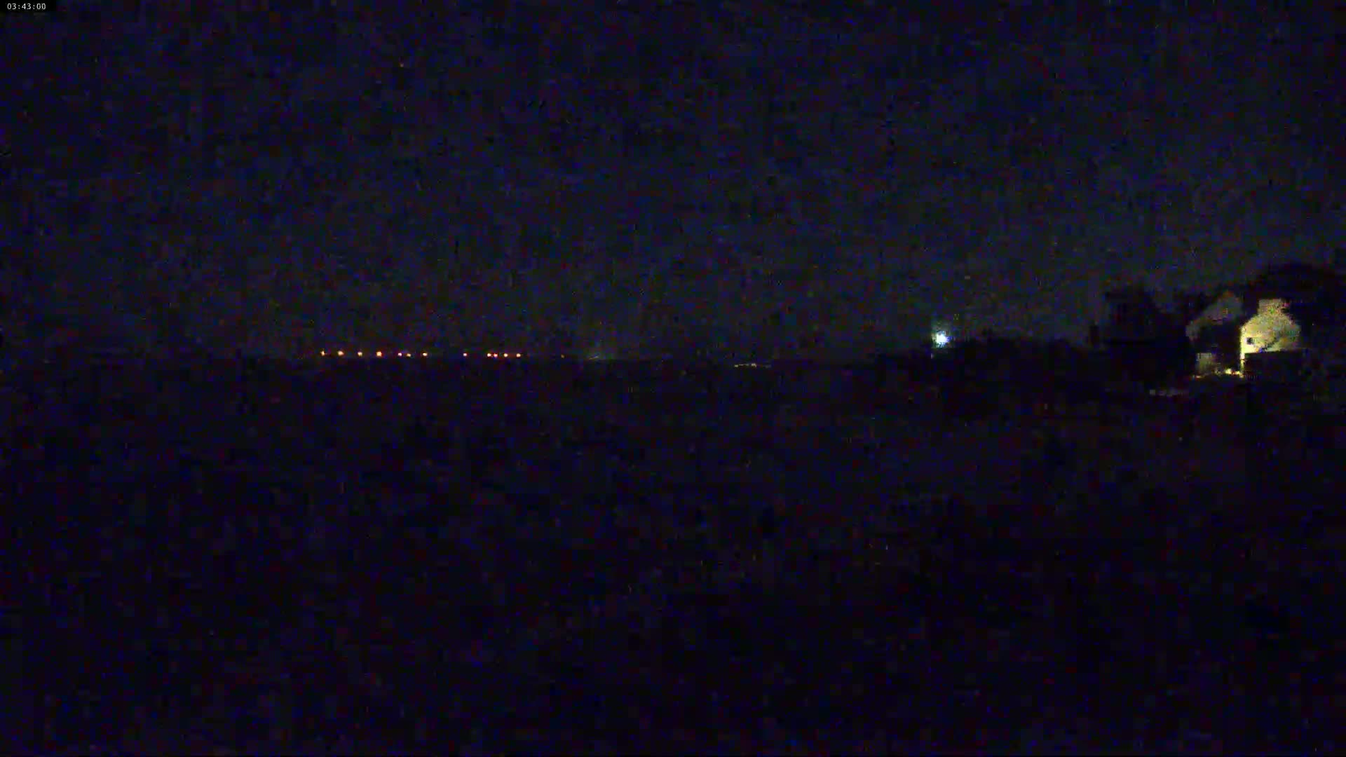 A dark, nighttime scene shows a line of distant lights and a dimly lit building in the foreground under a clear, starlit sky.
