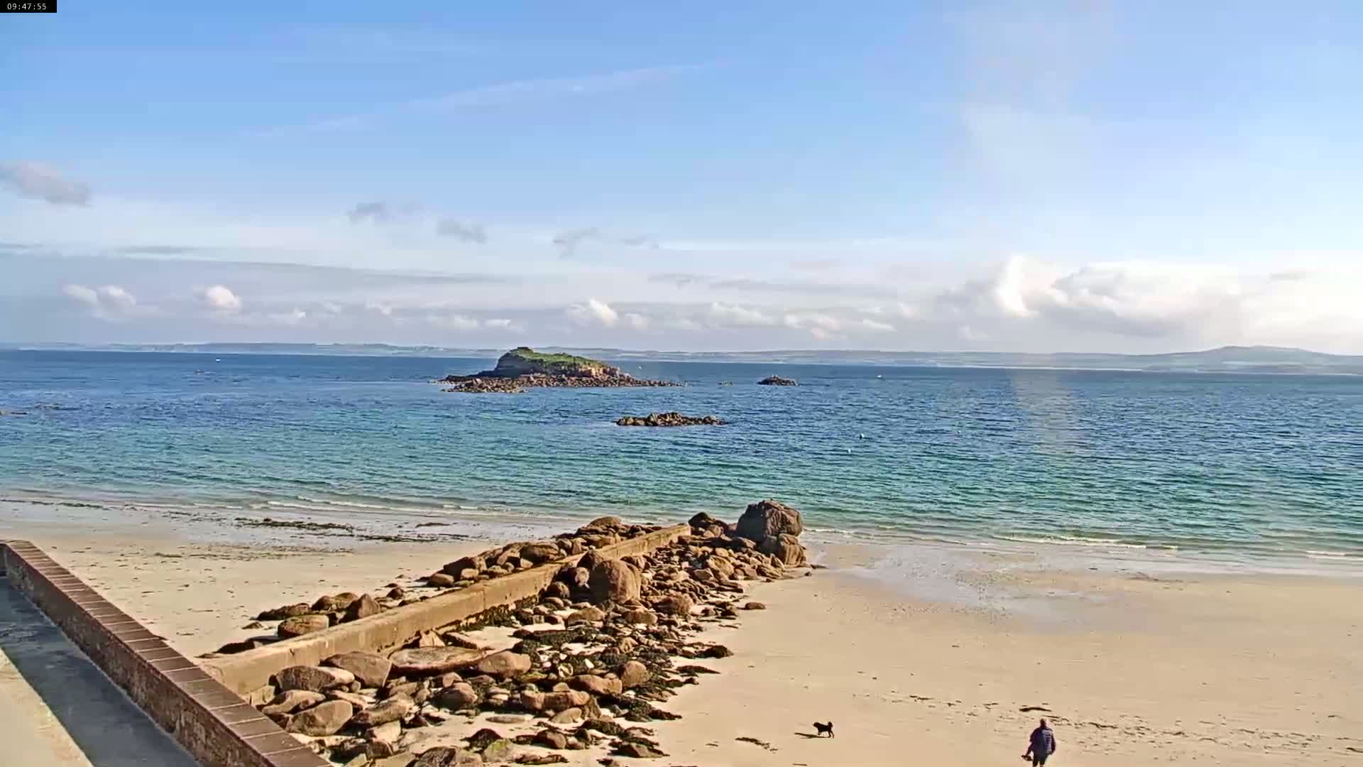 A sandy beach with a rocky jetty extends into a calm, turquoise ocean, under a mostly sunny sky, with small islands visible in the distance.