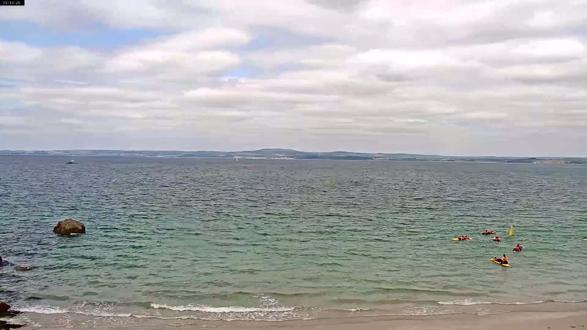 Several people are kayaking in a calm, light-green sea under a cloudy sky near a sandy beach.