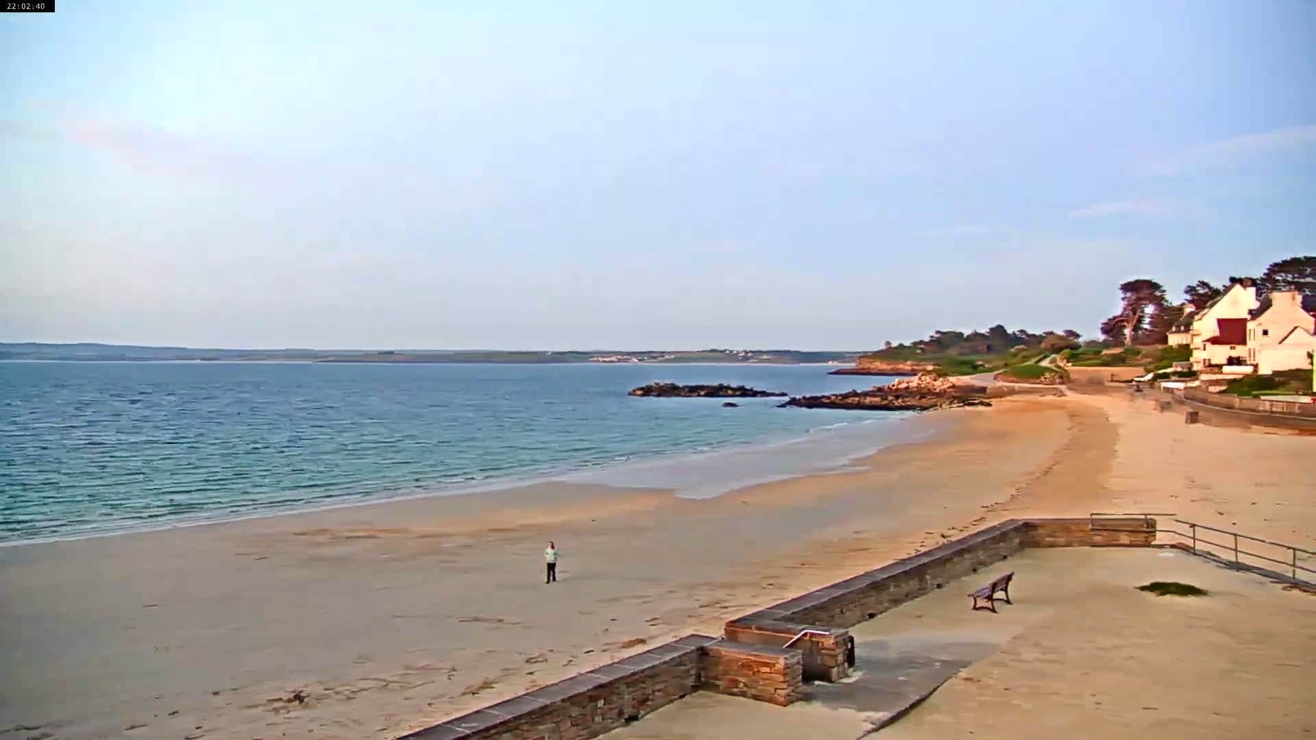 A calm, light-blue ocean meets a sandy beach where a lone person stands near a stone wall and a bench, with houses and trees visible in the distance under a clear sky.