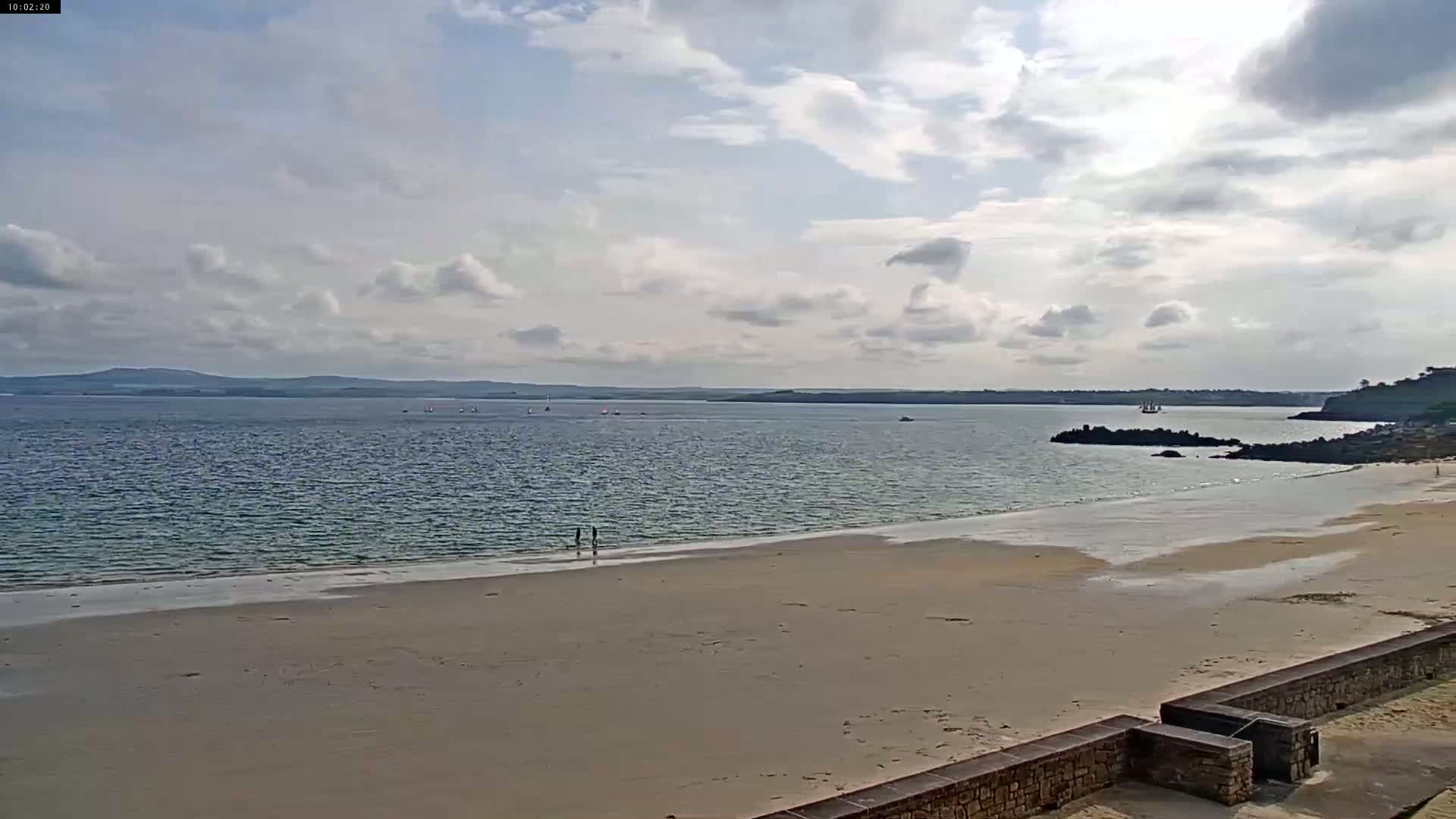 A sandy beach stretches towards a calm body of water under a partly cloudy sky, with a few small boats visible in the distance.