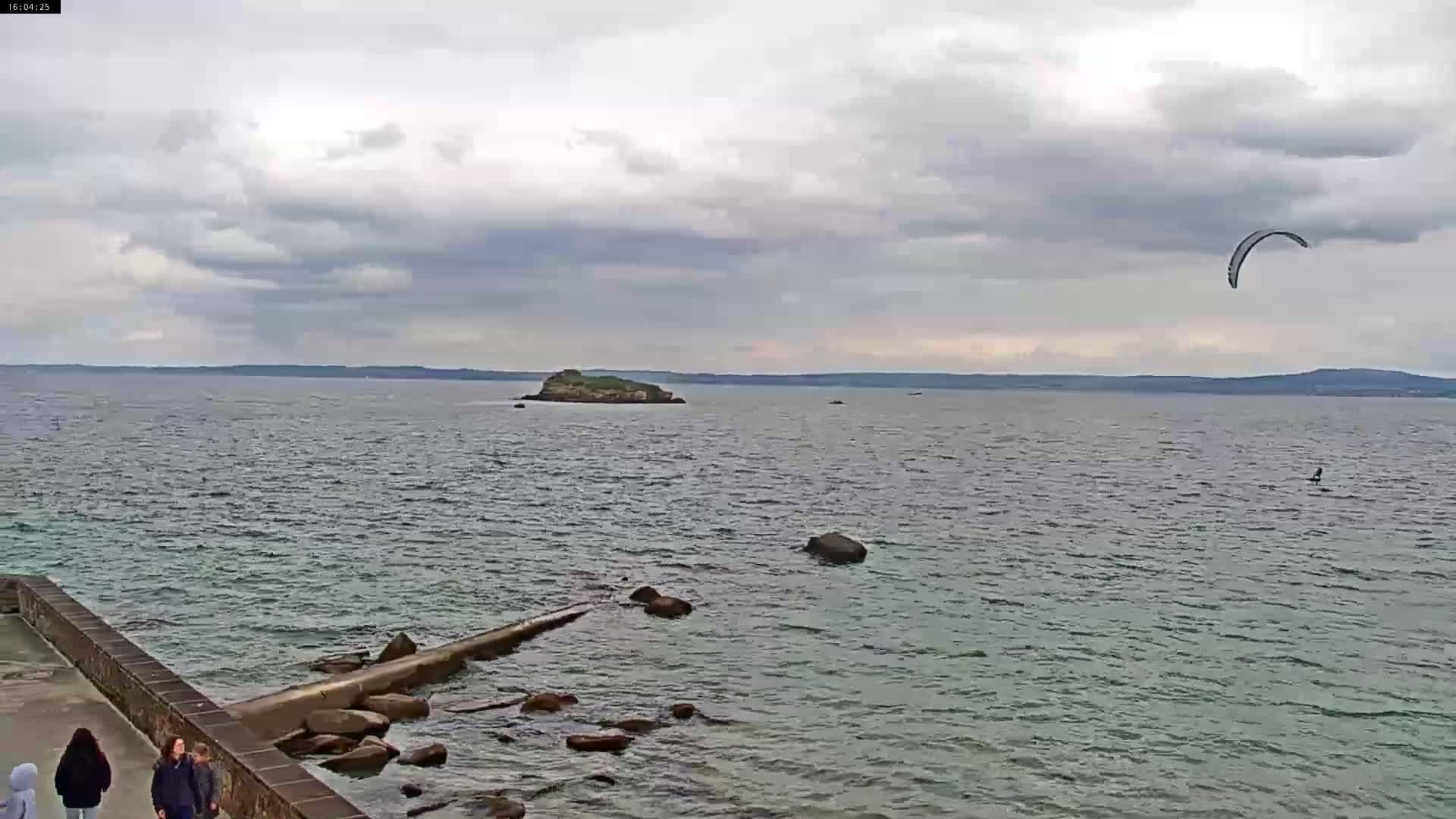 A body of water with a small island in the distance, a kiteboarder in the water, and a few people walking along a stone wall on the shore under an overcast sky.