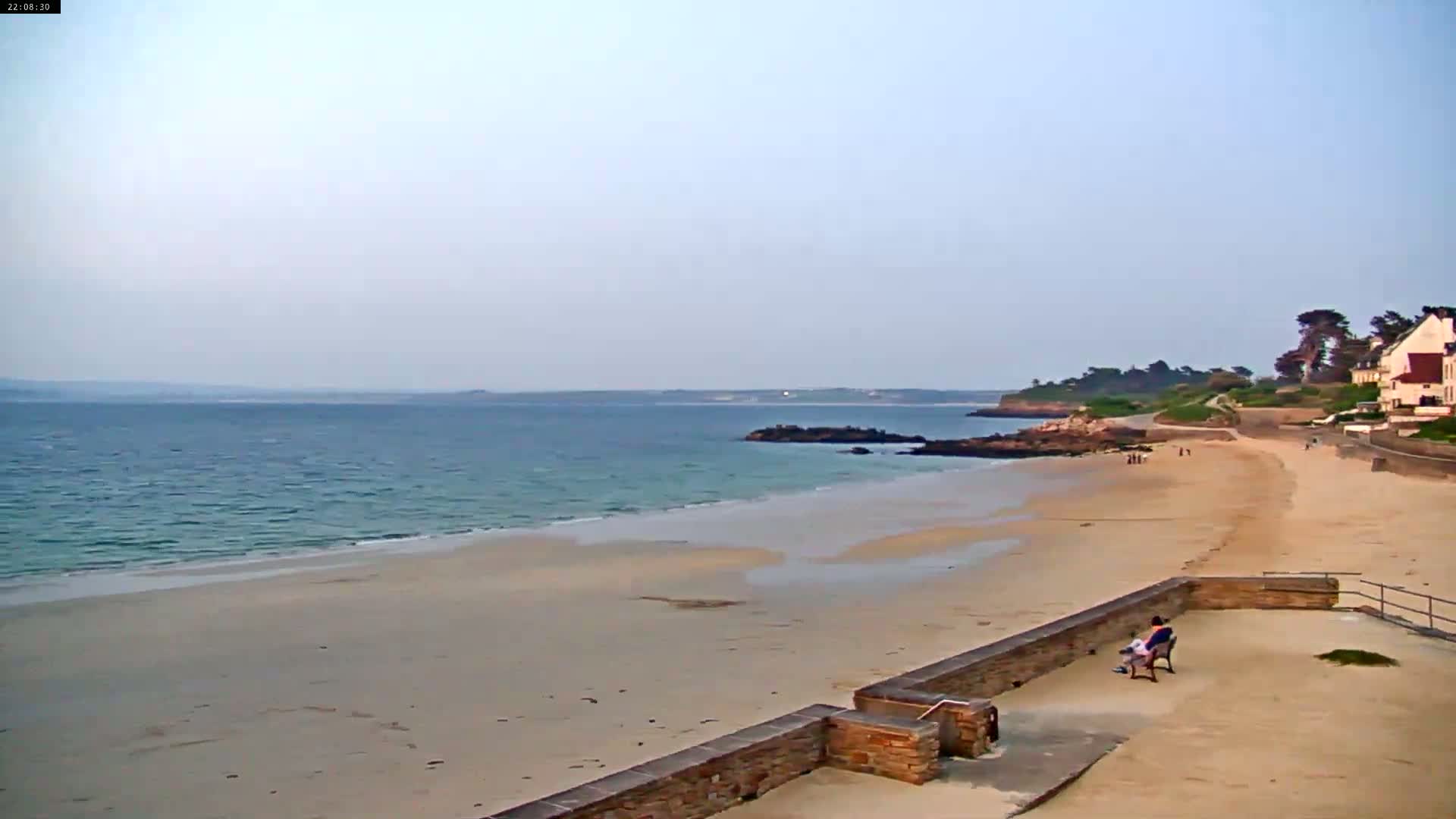 A person sits on a bench overlooking a calm, sandy beach that stretches to a rocky shoreline and calm ocean under a clear sky.