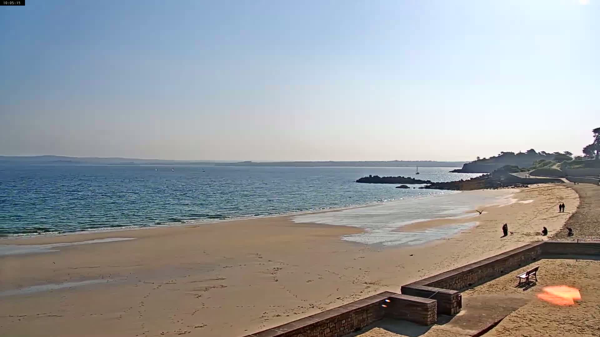 A sandy beach stretches along a calm, blue ocean under a clear, sunny sky; a few people are visible strolling along the shore.