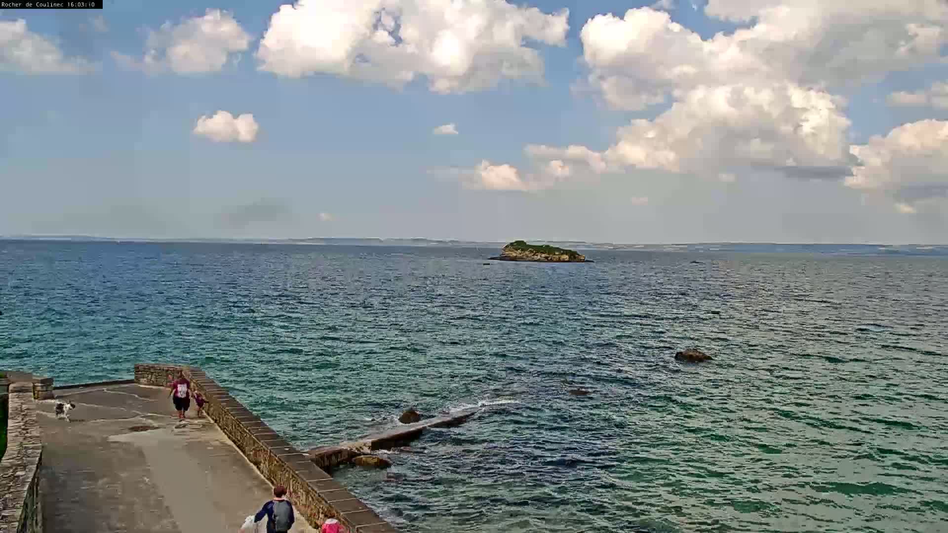 A partly cloudy day shows a calm ocean with a small island in the distance, as people walk along a stone path leading to the water.