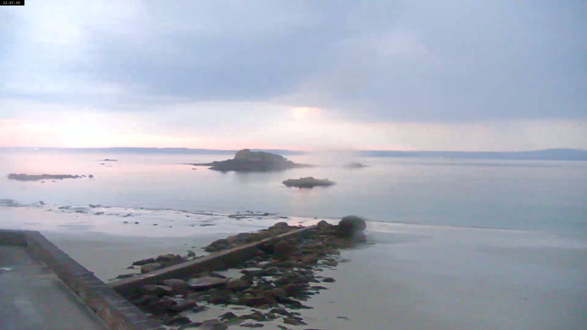 A rocky shoreline with a low wall leads to calm, light-grey water with small islands under a hazy, pale sky.
