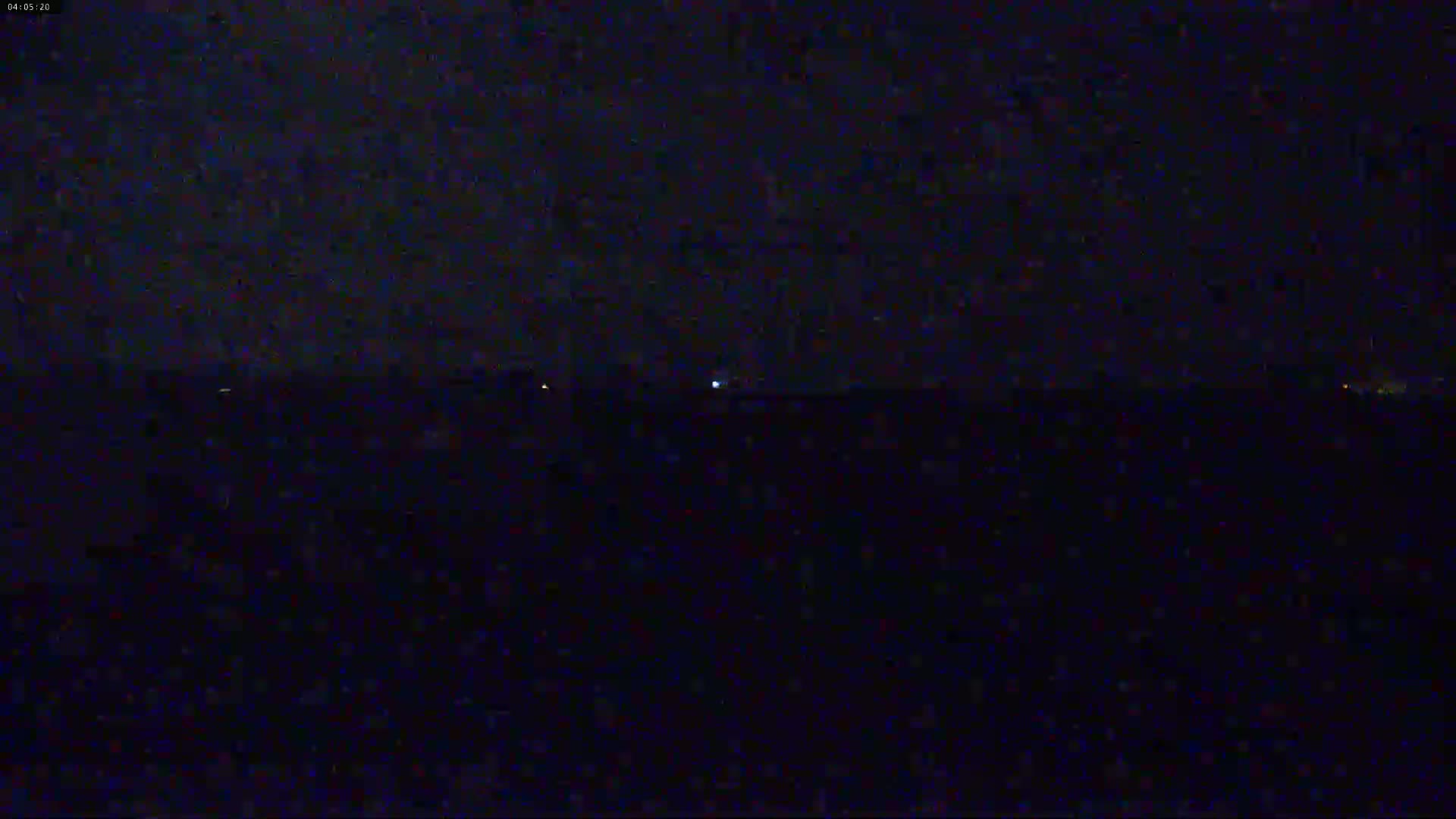 A dark, nighttime scene shows a flat, dark expanse with a few faint lights visible on the horizon under a mostly clear, dark sky.