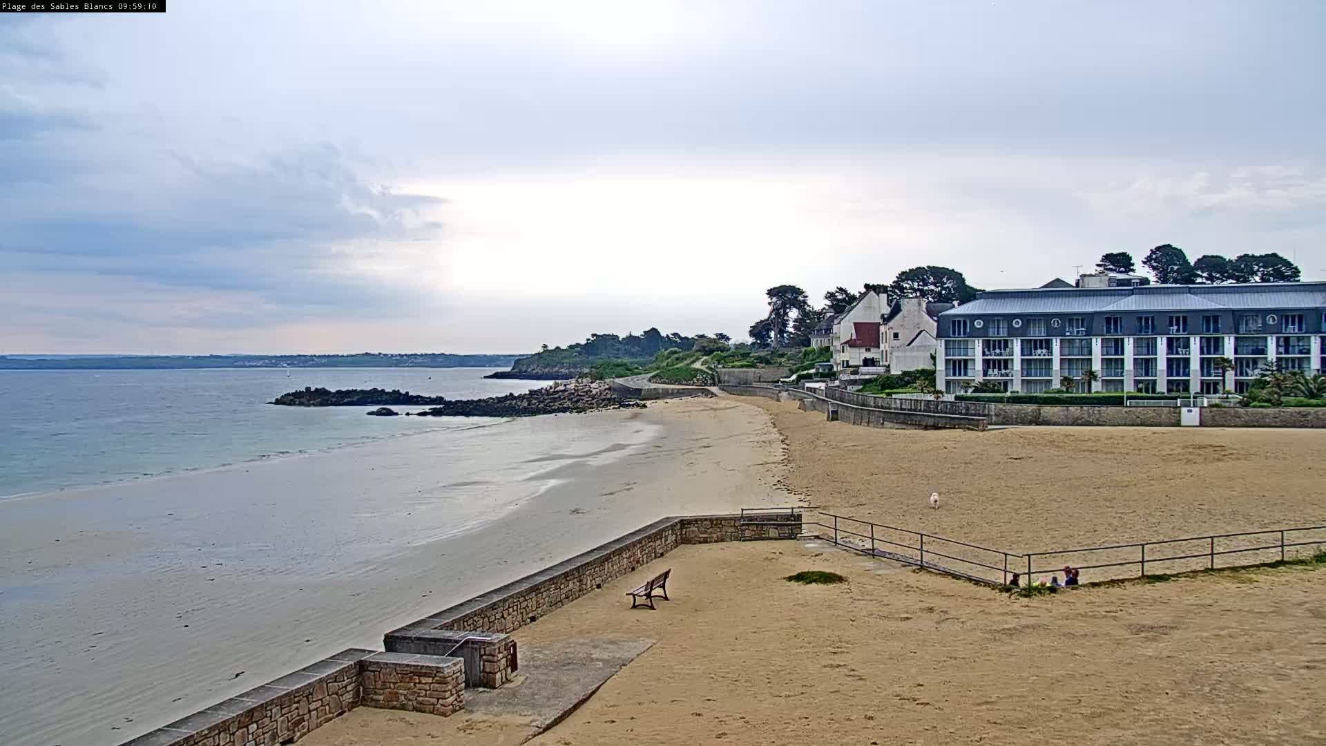 A sandy beach stretches alongside a calm ocean under a cloudy sky, with a multi-story building and a few people visible in the background.
