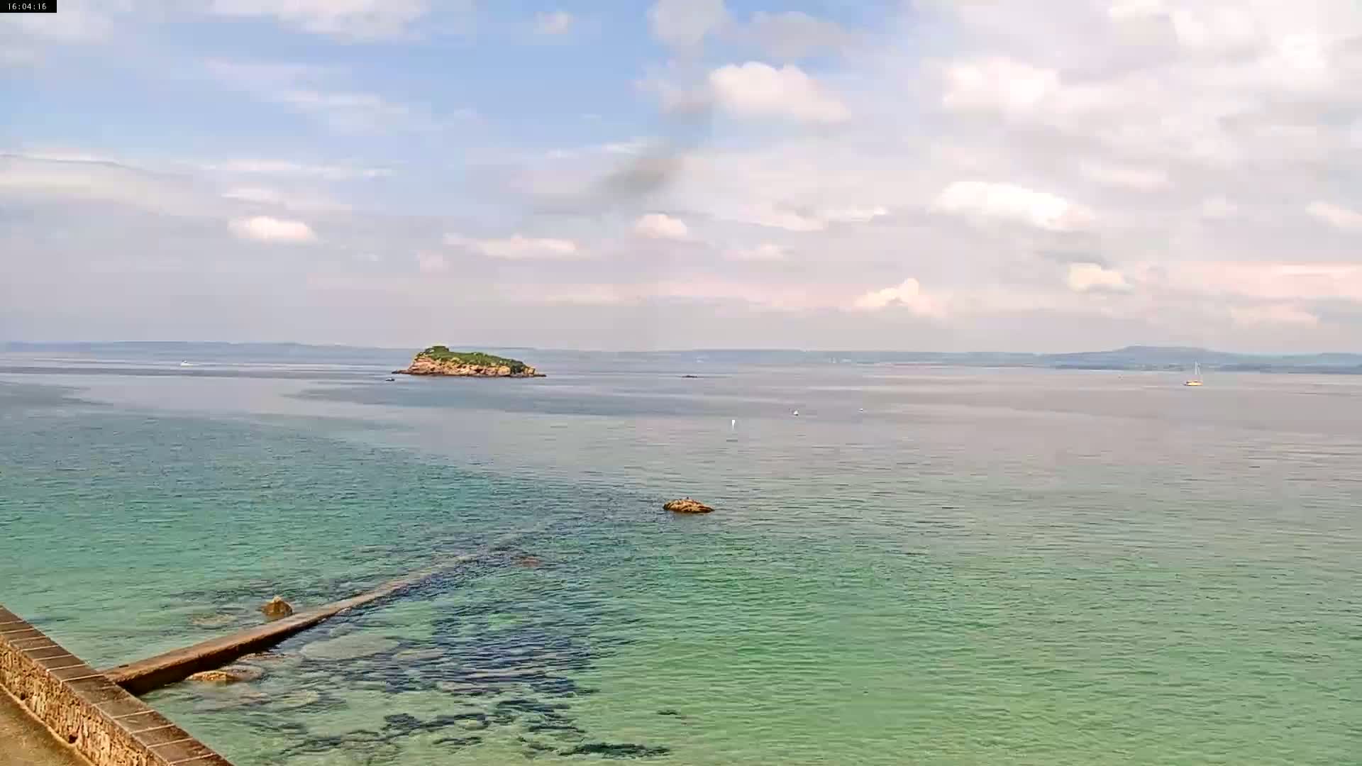 A partly cloudy day reveals a tranquil coastal scene with a small island in a calm, teal-colored sea, a stone jetty extending into the water, and a distant shoreline.