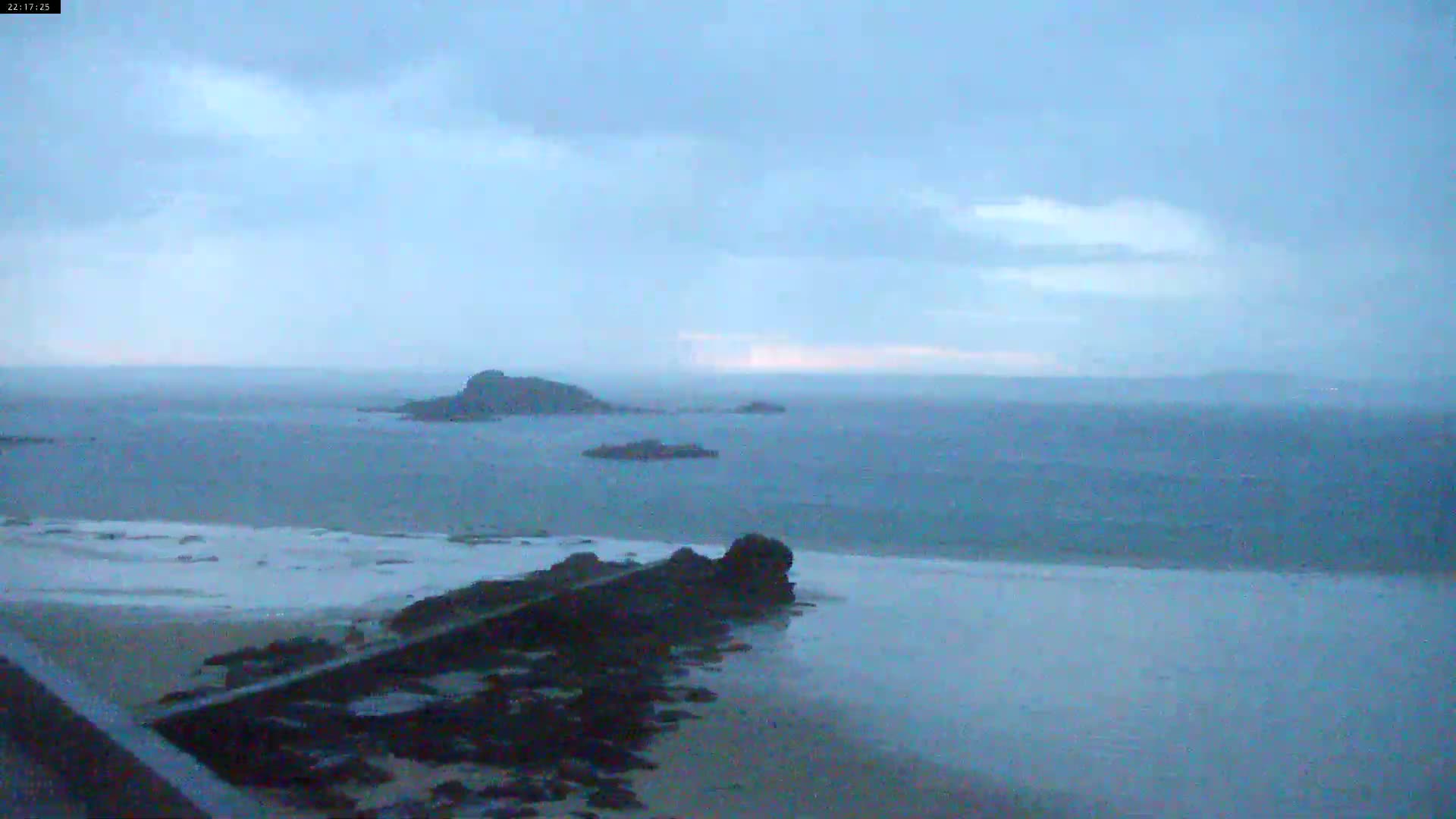 A calm, overcast day reveals a sandy beach, dark rocks, and several small islands in the distance, across a body of water.