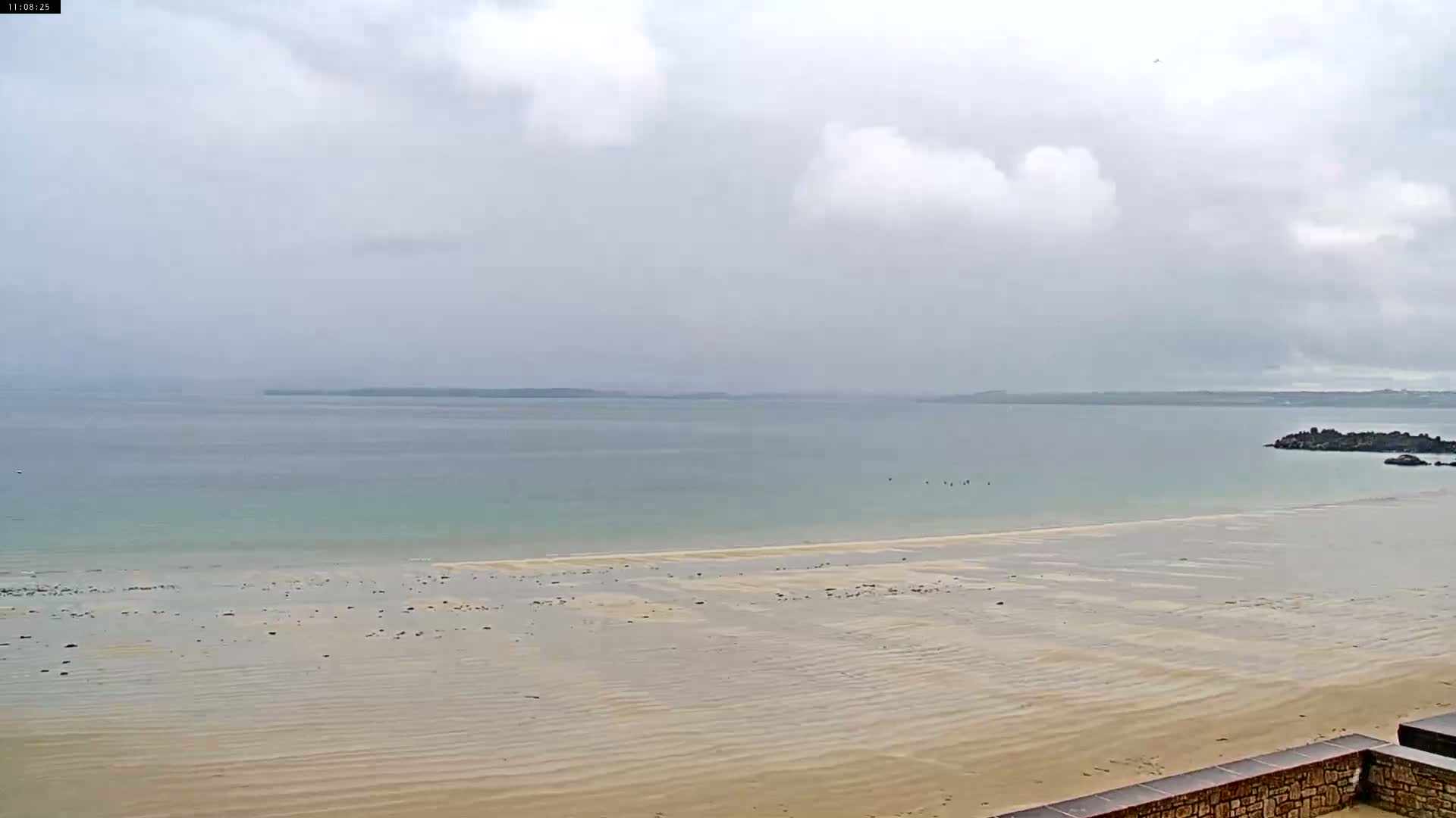 A wide sandy beach stretches to a calm, light-green ocean under a cloudy sky.