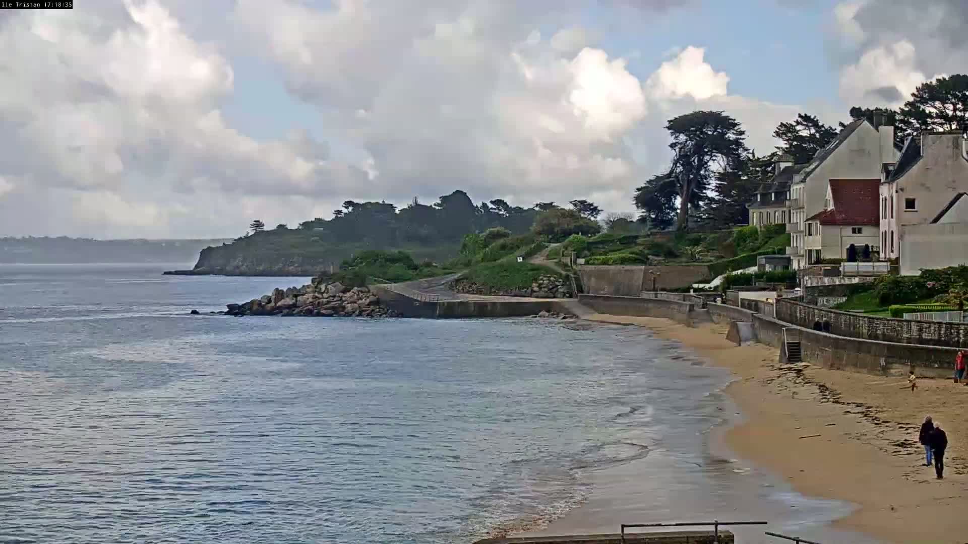 A sandy beach with gentle waves laps a shoreline where several houses sit atop a low cliff under a partly cloudy sky.