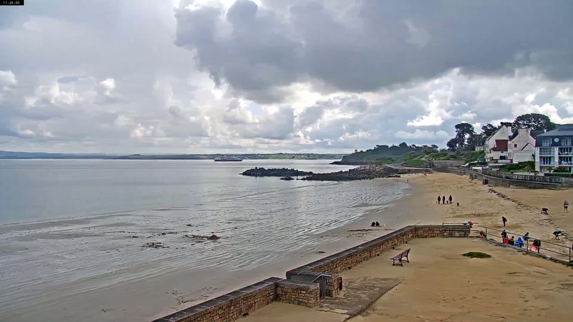 A sandy beach with a few people on it stretches out to a calm bay under a partly cloudy sky.