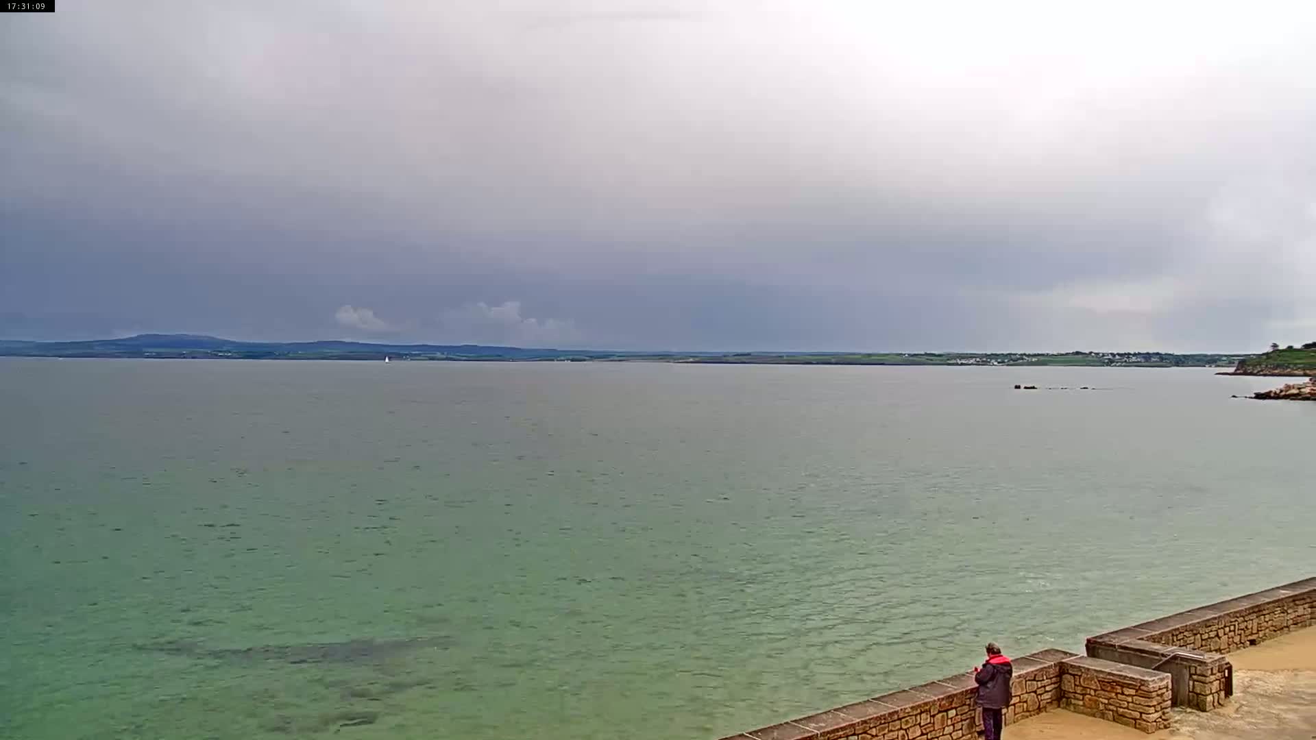 A person stands on a stone wall overlooking a calm, greenish-grey sea under an overcast sky.